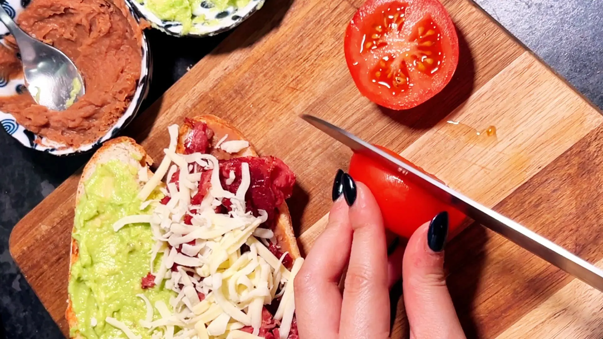 A hand with black nail polish slices a tomato next to a baguette topped with avocado, shredded cheese, and meat.
