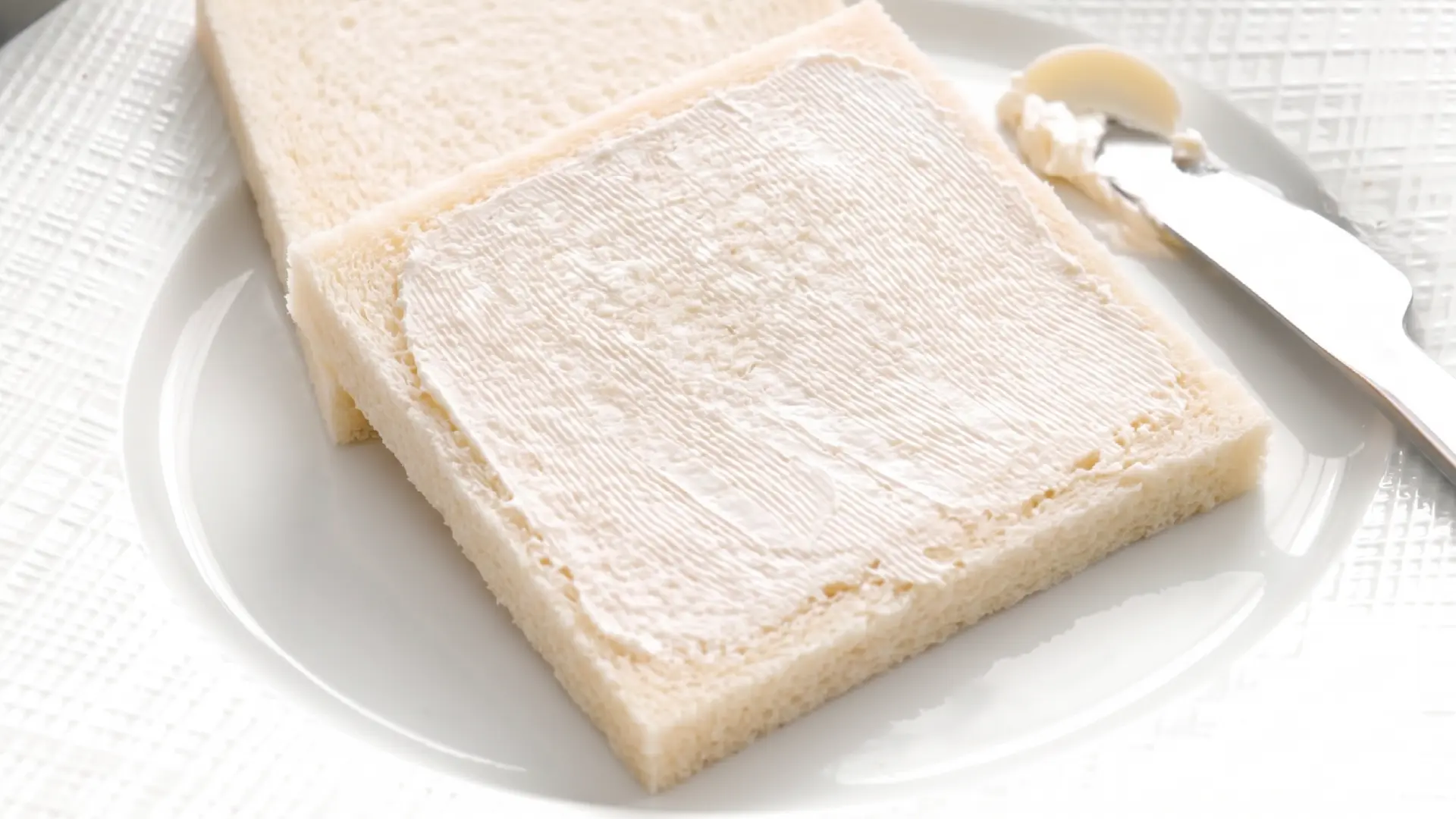 A slice of flattened crustless white bread spread with cream cheese on a white plate next to a butter knife