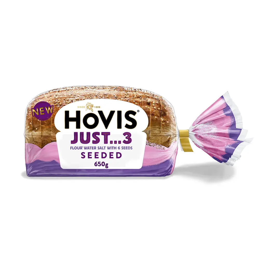 A loaf of Hovis Just 3 seeded bread in clear and purple packaging with a yellow tie clip, set against a plain background.