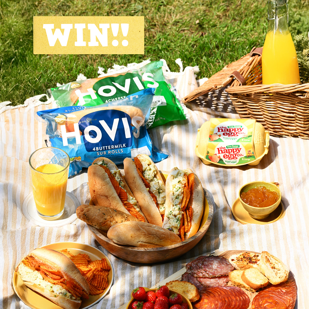 A vibrant top-down shot of a picnic spread on green grass, featuring a "WIN!!" text overlay in the top left. The picnic includes two large bags of Hovis sub rolls, two yellow cartons of The Happy Egg Co. eggs, and a picnic basket. A wooden bowl holds several filled sub rolls, likely egg salad sandwiches, while another plate has a partially eaten sub and wavy crisps. Other food items visible are a bowl of fresh strawberries, a platter of sliced cured meats, and a small dish of jam or chutney. Two glasses of orange juice, with a bottle in the background, complete the sunny outdoor scene.