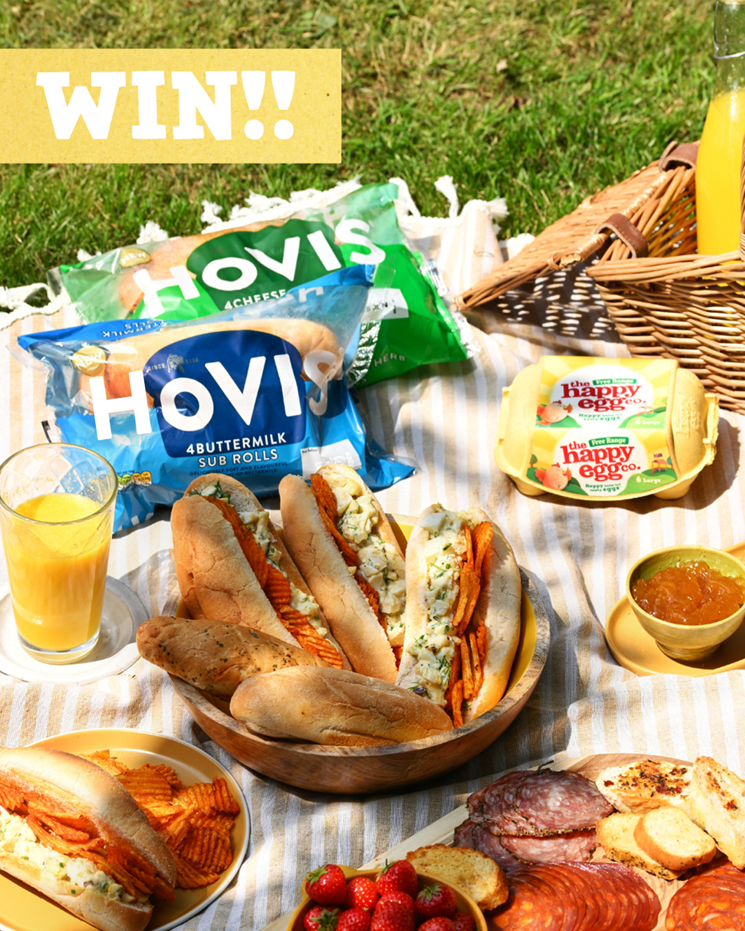 A vibrant top-down shot of a picnic spread on green grass, featuring a "WIN!!" text overlay in the top left. The picnic includes two large bags of Hovis sub rolls, two yellow cartons of The Happy Egg Co. eggs, and a picnic basket. A wooden bowl holds several filled sub rolls, likely egg salad sandwiches, while another plate has a partially eaten sub and wavy crisps. Other food items visible are a bowl of fresh strawberries, a platter of sliced cured meats, and a small dish of jam or chutney. Two glasses of orange juice, with a bottle in the background, complete the sunny outdoor scene.