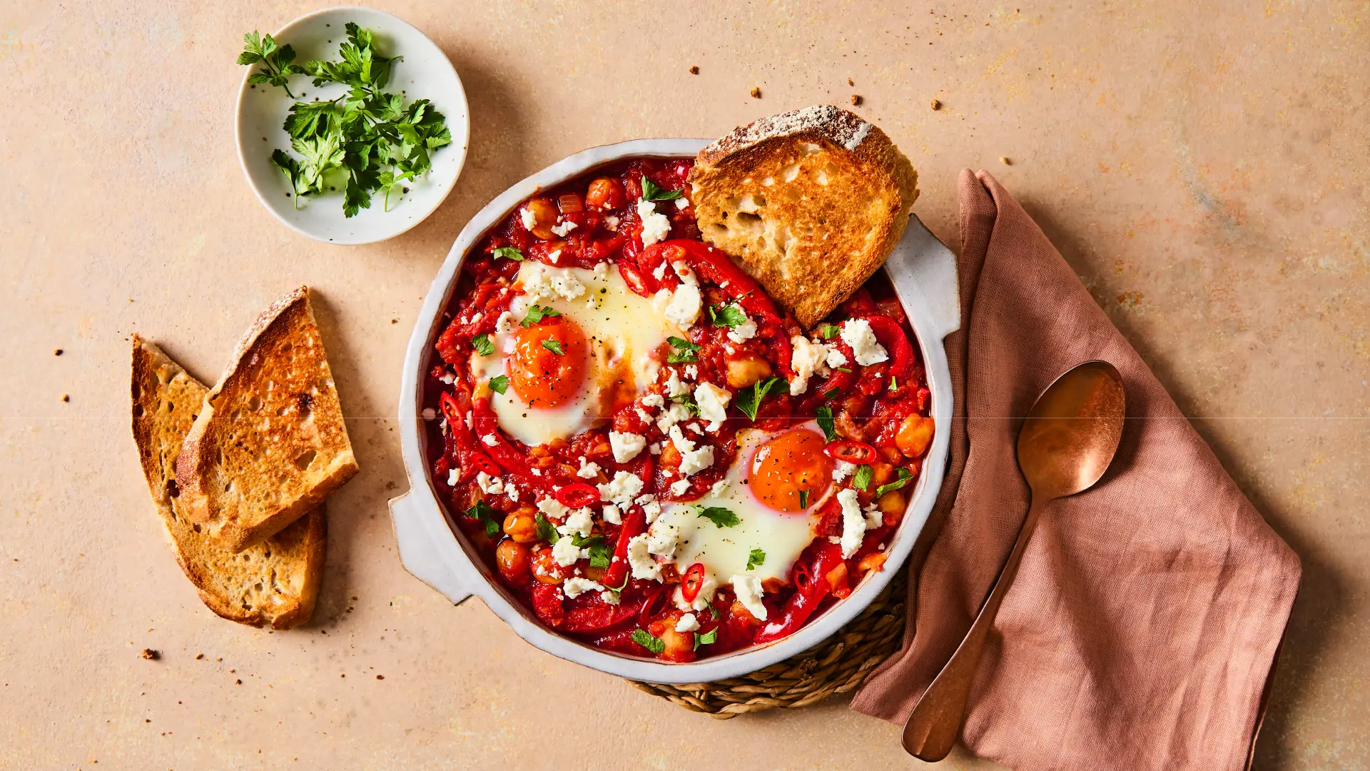 An overhead food photography shot of a single serving of Shakshuka, a dish of eggs poached in a flavourful tomato and pepper sauce, served in a round, shallow, off-white stoneware dish. The dish includes visible chickpeas, red peppers, and is generously topped with crumbled white feta cheese. Two eggs with bright orange yolks are visible near the centre. It is garnished with fresh parsley on the side and served with two slices of grilled or toasted bread. A copper spoon rests on a folded peach-coloured linen napkin beside the dish, all set on a light tan surface.