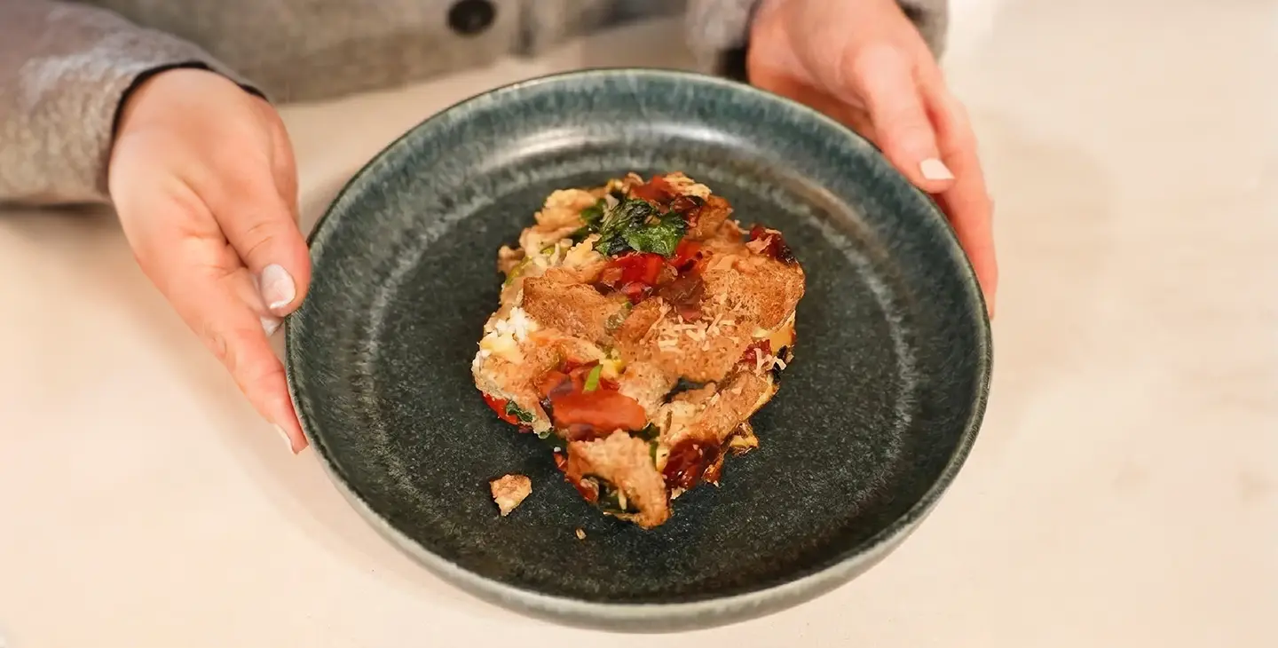 A person's hands hold a dark ceramic plate with a square serving of savoury vegetable bread pudding or strata.
