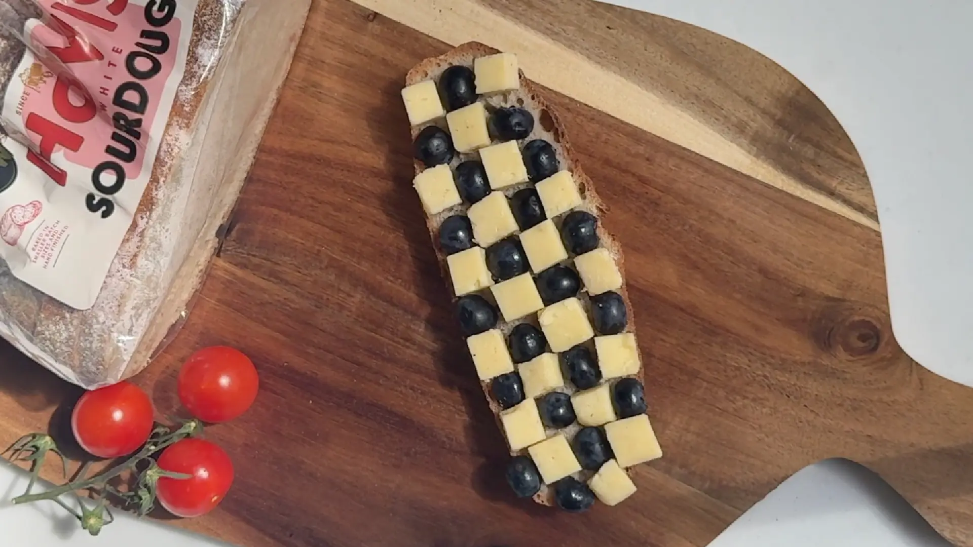 A slice of sourdough toast with a checkerboard pattern of cheese cubes and blueberries on a wooden board.