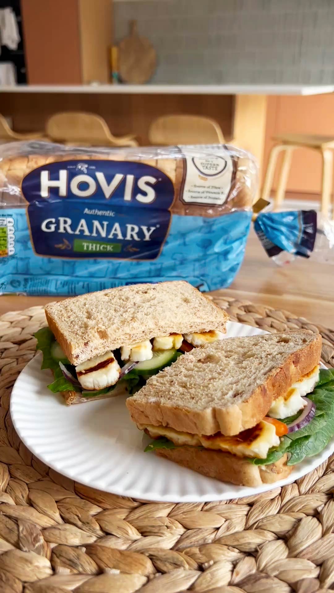 A sandwich cut in half sitting on a white plate, presented on a woven placemat. Hovis Granary in the background.