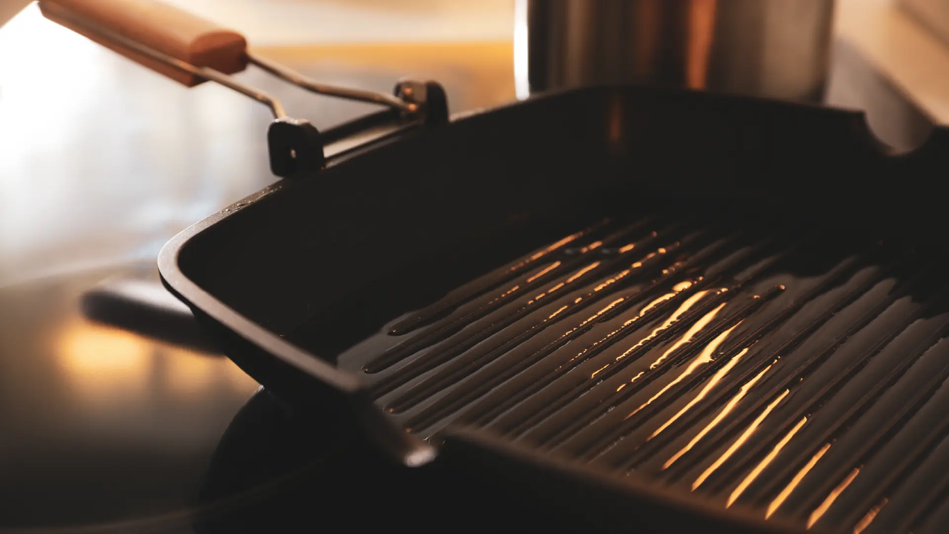 Close up of a black ribbed grill pan with cooking oil, sitting on a stovetop with warm light reflecting off the surface.
