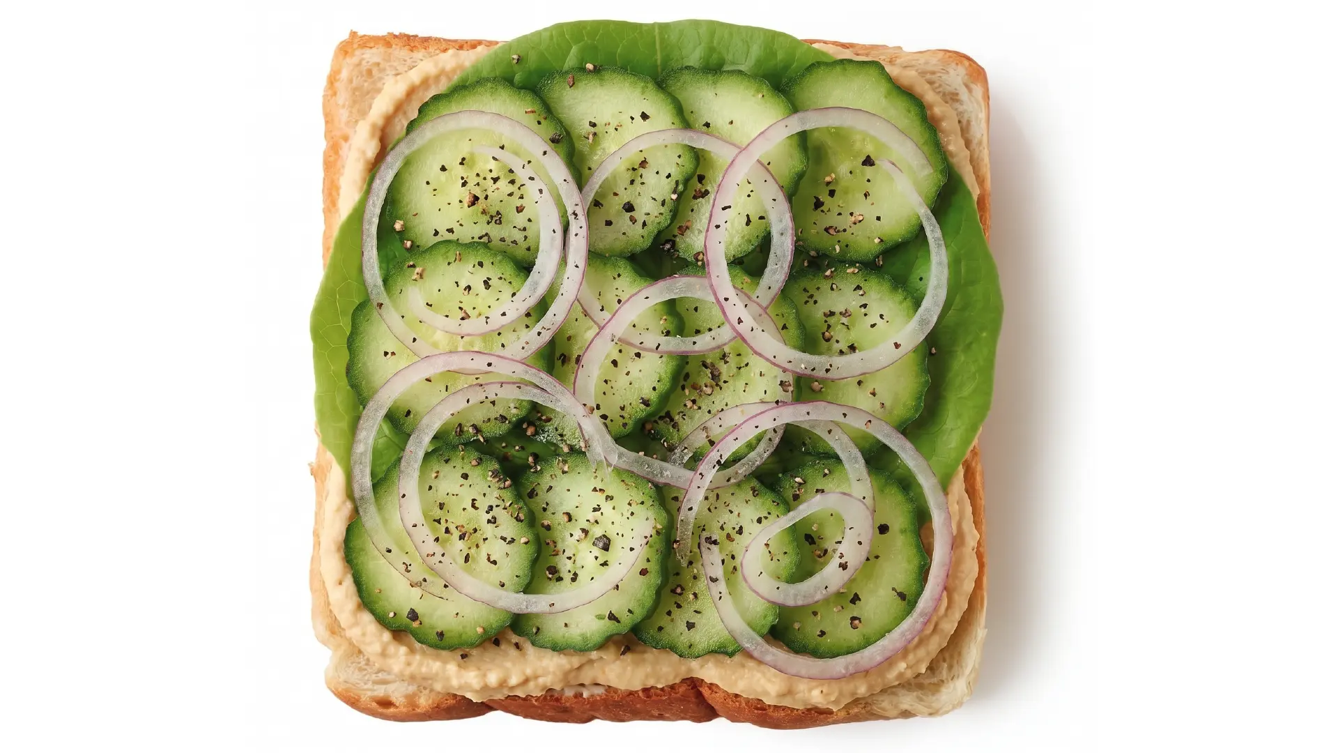 Top down view of open faced sandwich topped with hummus, lettuce, cucumber, thinly sliced red onion and black pepper