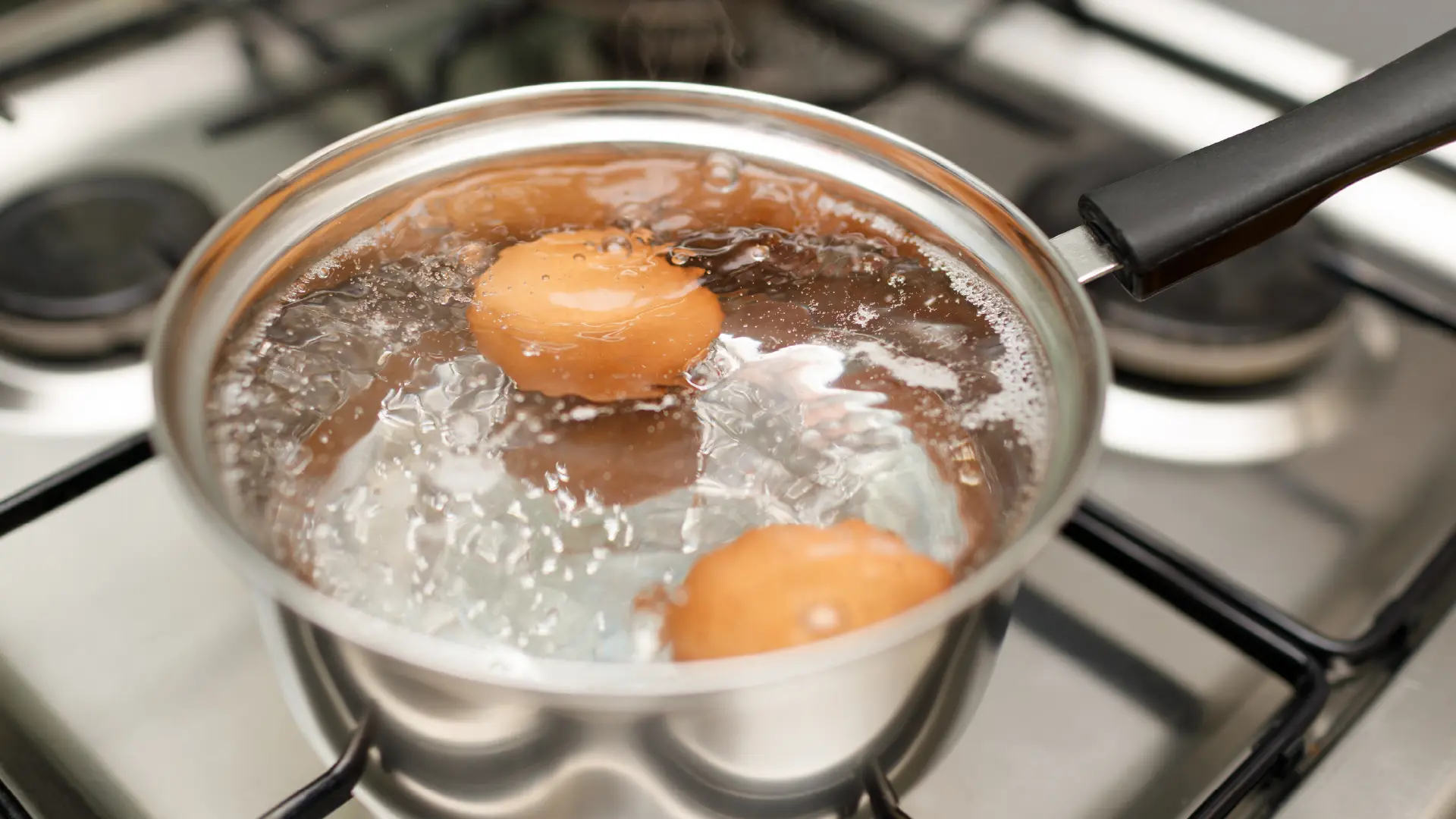 Two whole brown eggs in a small metal saucepan with vigorously boiling water, sitting on a lit gas stove burner.