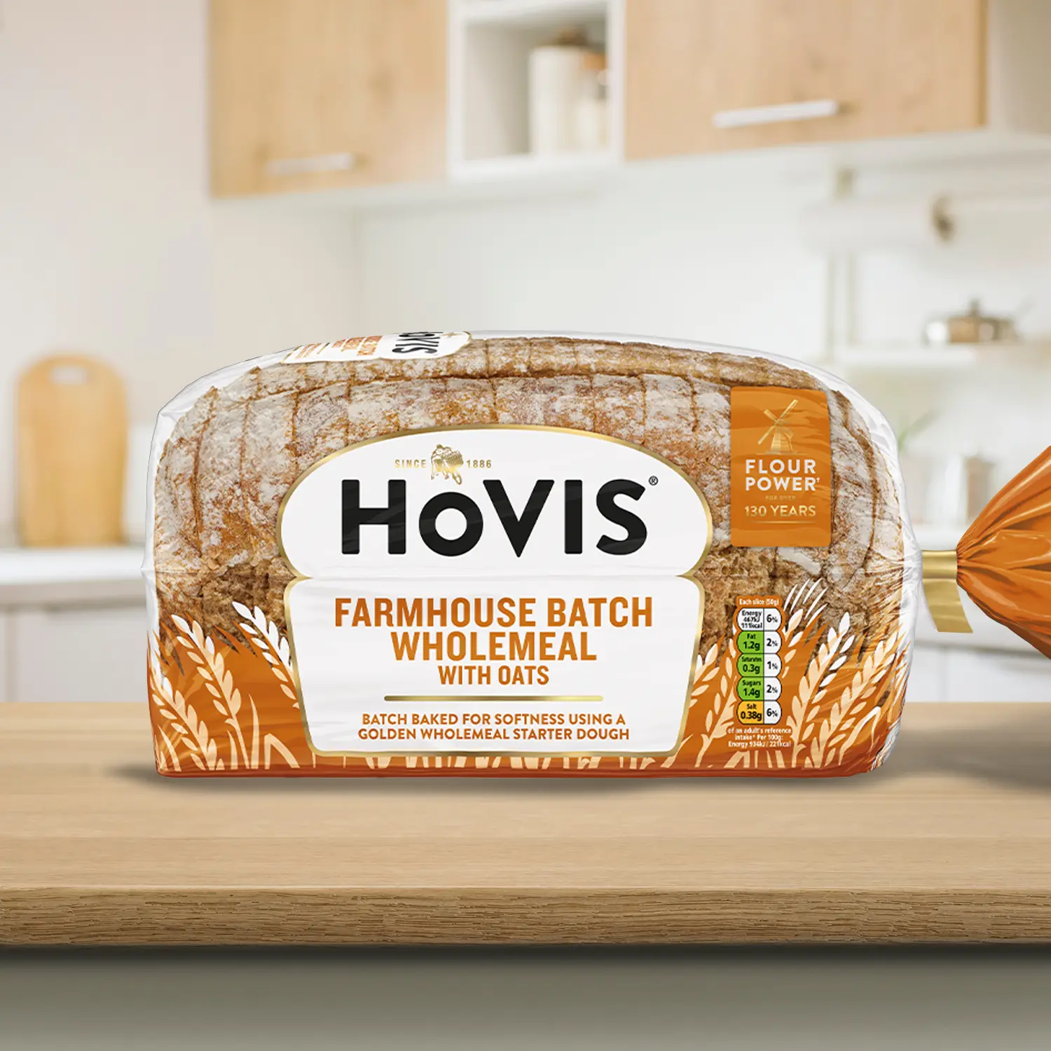A loaf of Farmhouse Batch Wholemeal with Oats bread with sourdough and a crust dusted with flour, displayed on a wooden kitchen countertop. The packaging features a white and orange design with a gold trim and nutrition information. 