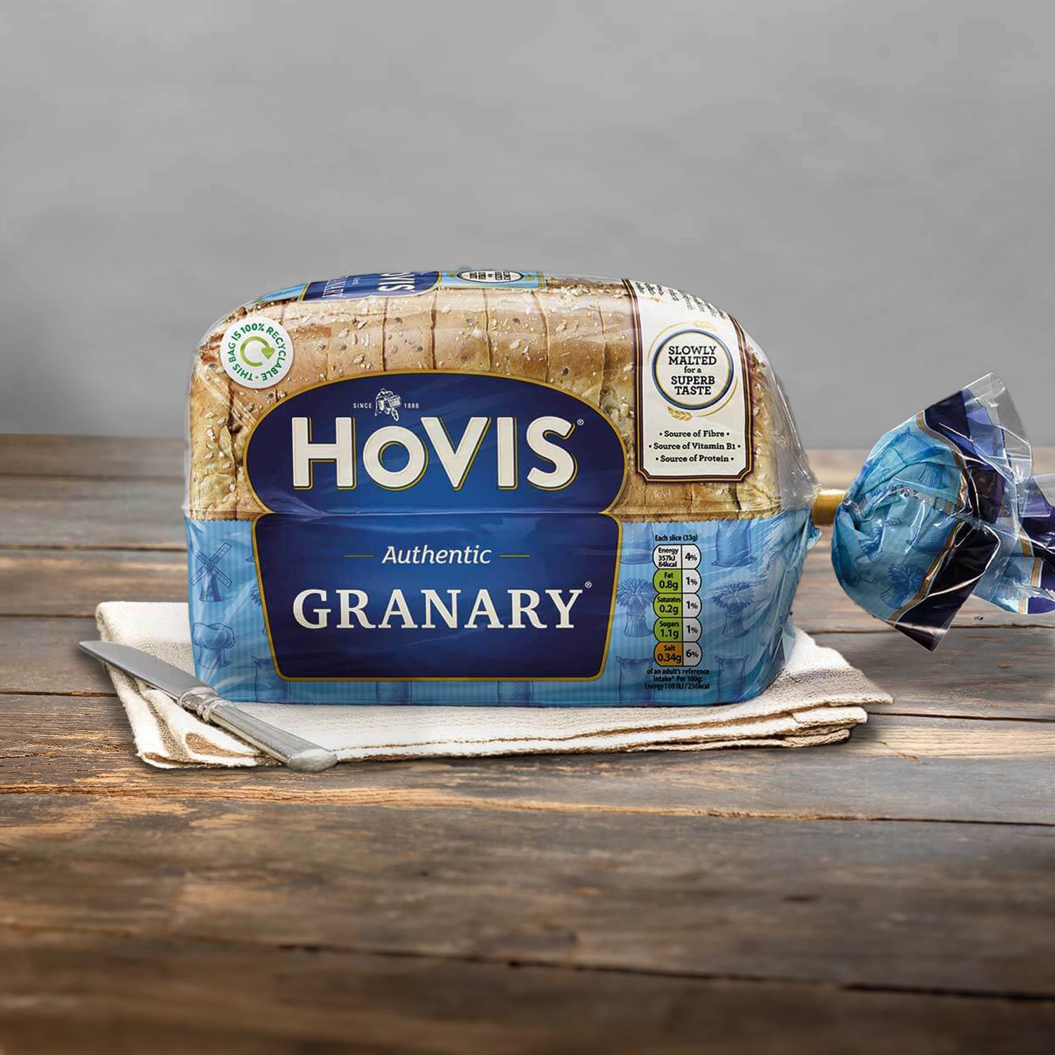 Hovis Granary 400g loaf on beige napkins against a light grey background, resting on a rustic wooden table with natural light.