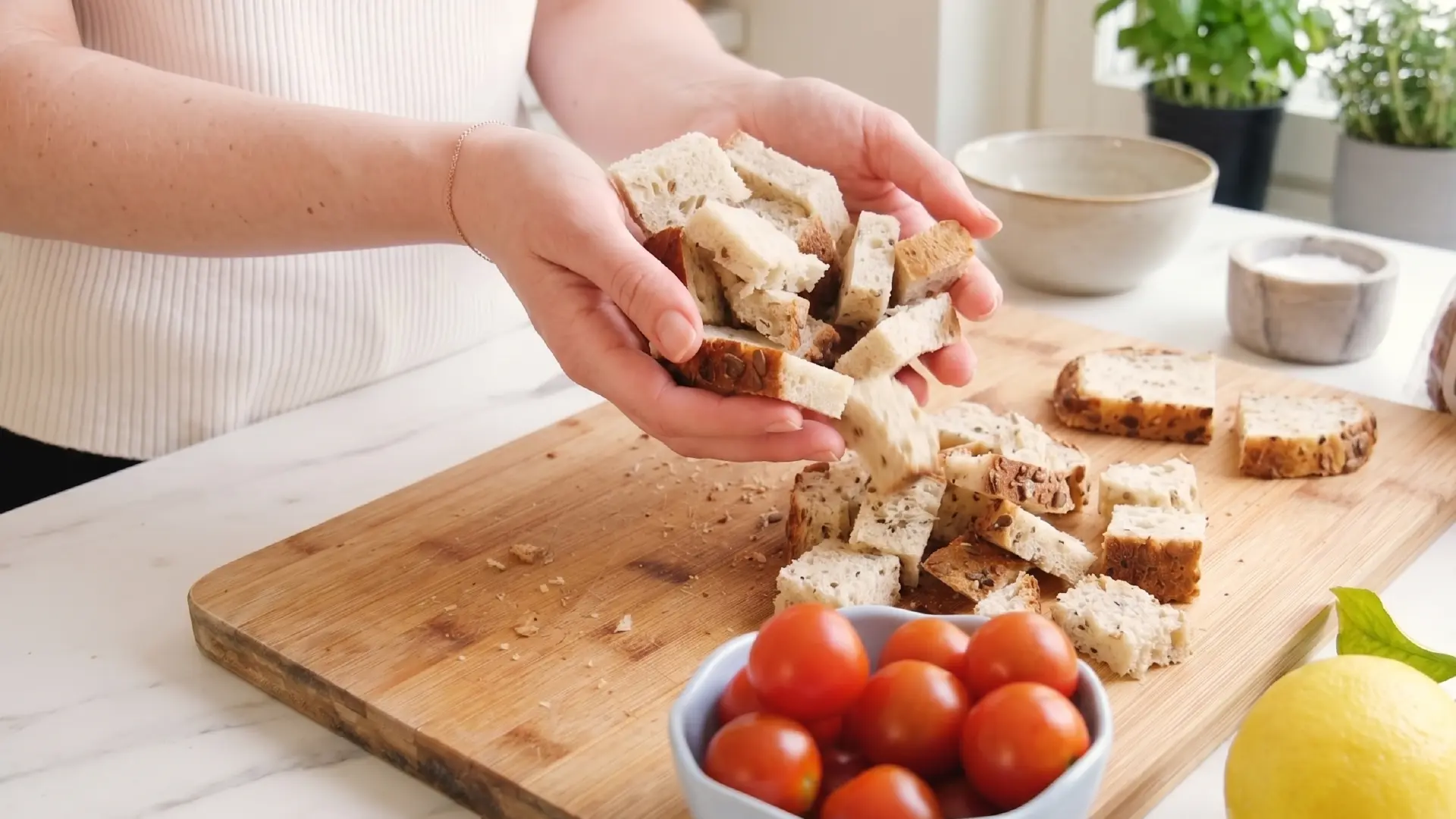 Hands holding cubed bread drop the pieces onto a wooden cutting board next to a small blue bowl of cherry tomatoes.