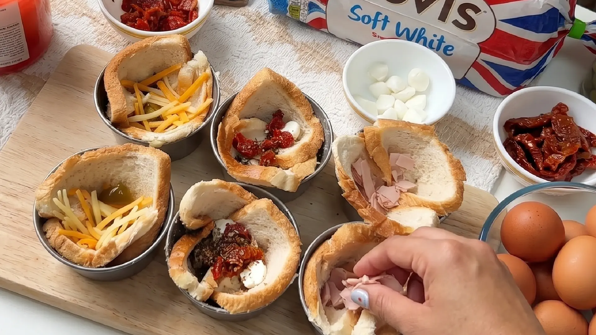 Bread slices in tins being filled with various ingredients like cheese, ham, and sun-dried tomatoes to make breakfast cups.