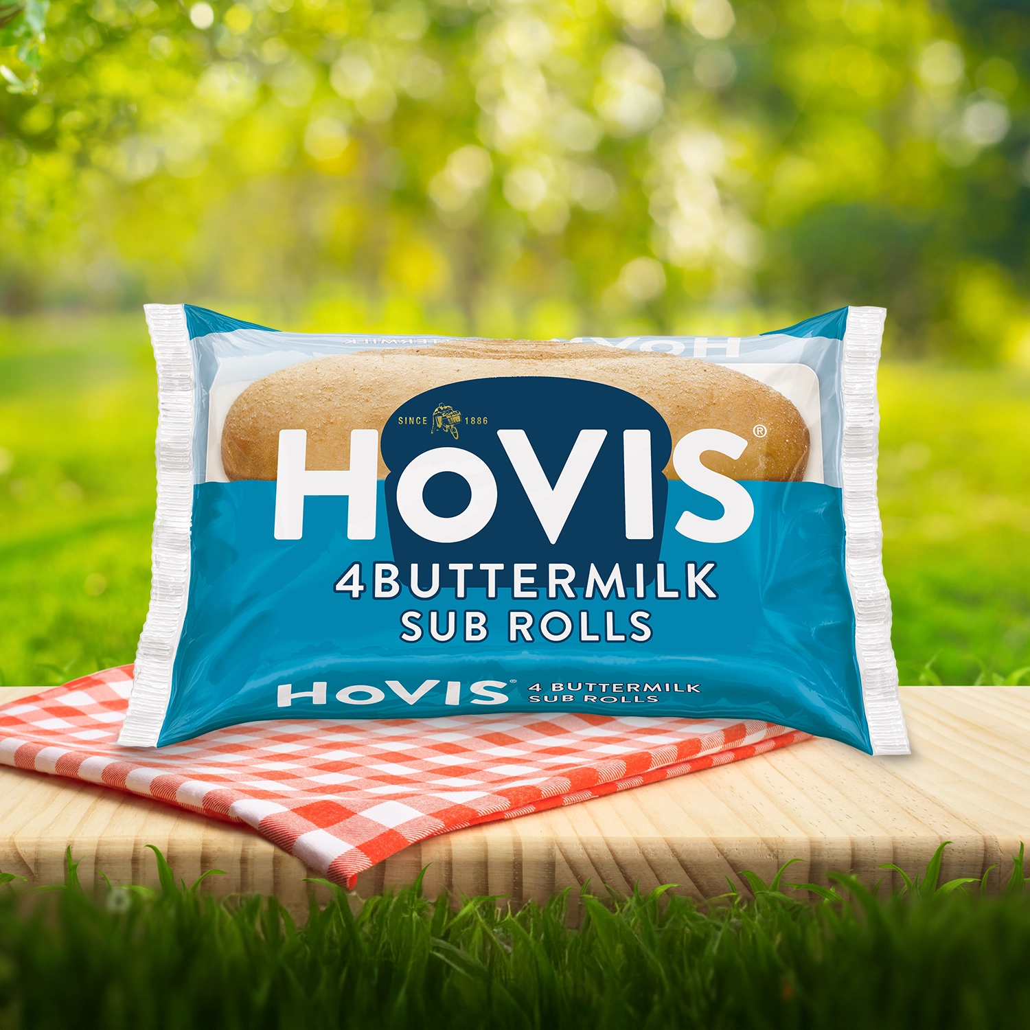 Hovis Buttermilk Sub Rolls packaging on a picnic table with a green outdoor background. The pack shows soft sub rolls  and highlights “Deliciously soft and flavourful with a hint of buttermilk.” A red gingham cloth adds a picnic feel to the setting.