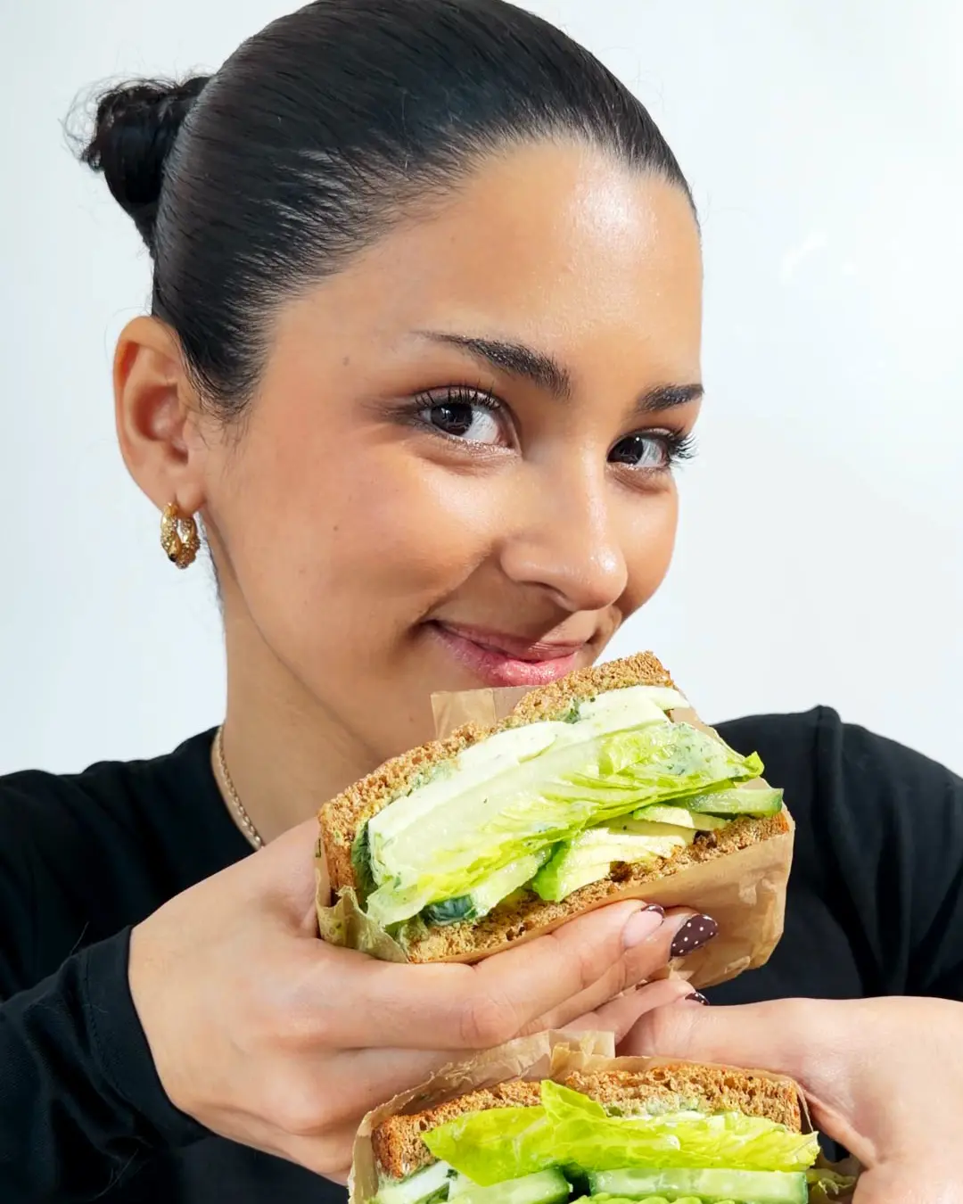 A woman smiling while holding a brown bread sandwich wrapped in paper, packed with fresh green lettuce and cucumber.