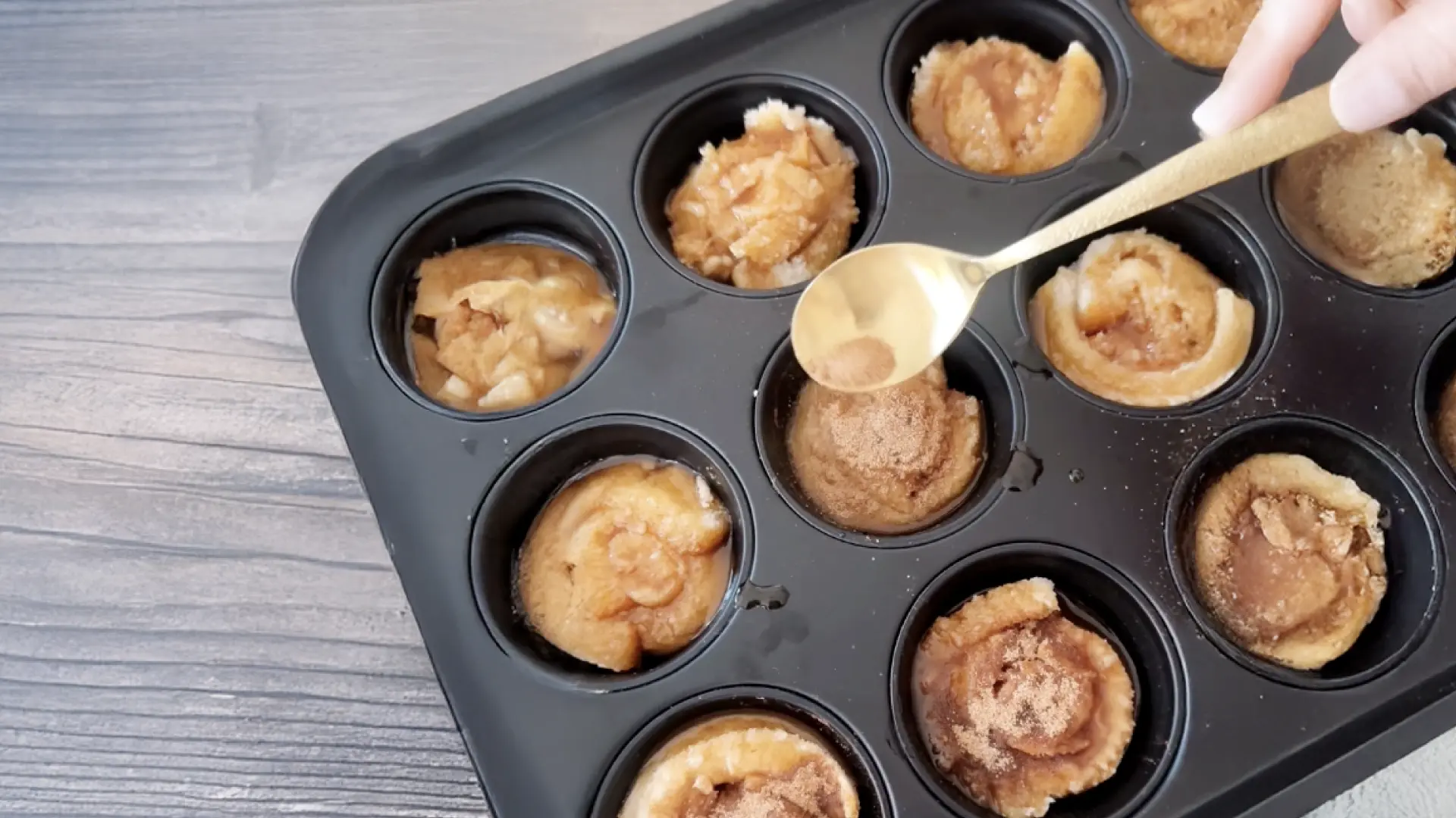A hand using a gold spoon to coat unbaked roll spirals in a black muffin tin with sweet liquid topping.