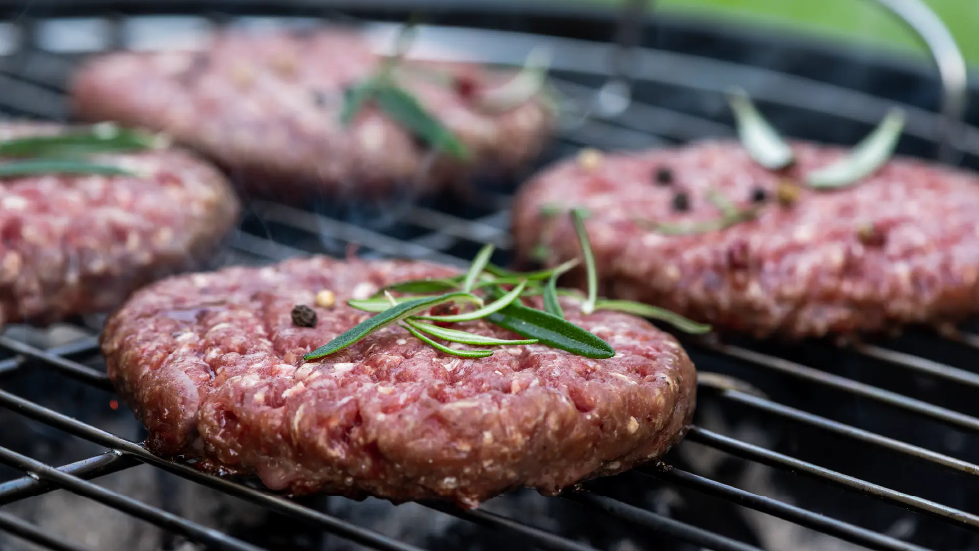 Four raw beef burger patties seasoned with peppercorns and fresh rosemary grilling on a metal barbecue grate.