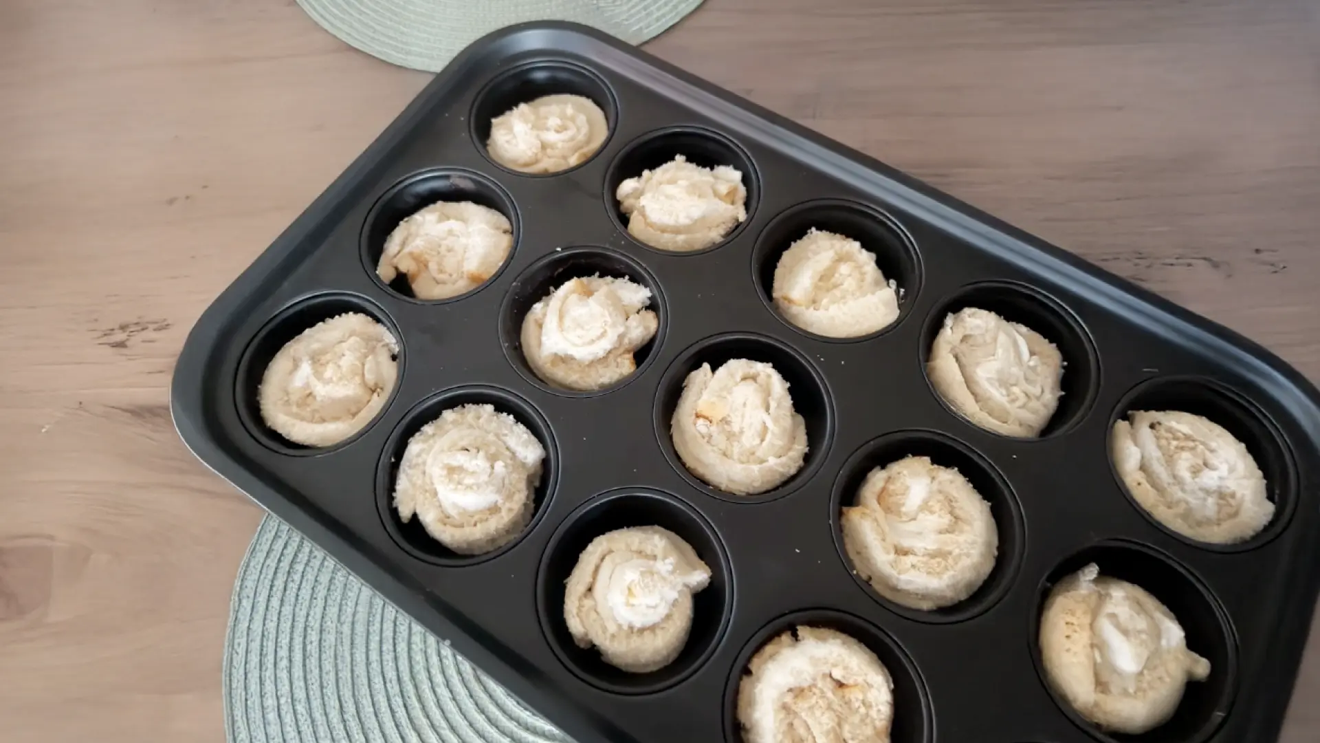 A black muffin tin filled with unbaked rolled bread spirals resting on a wooden table with a light green placemat.