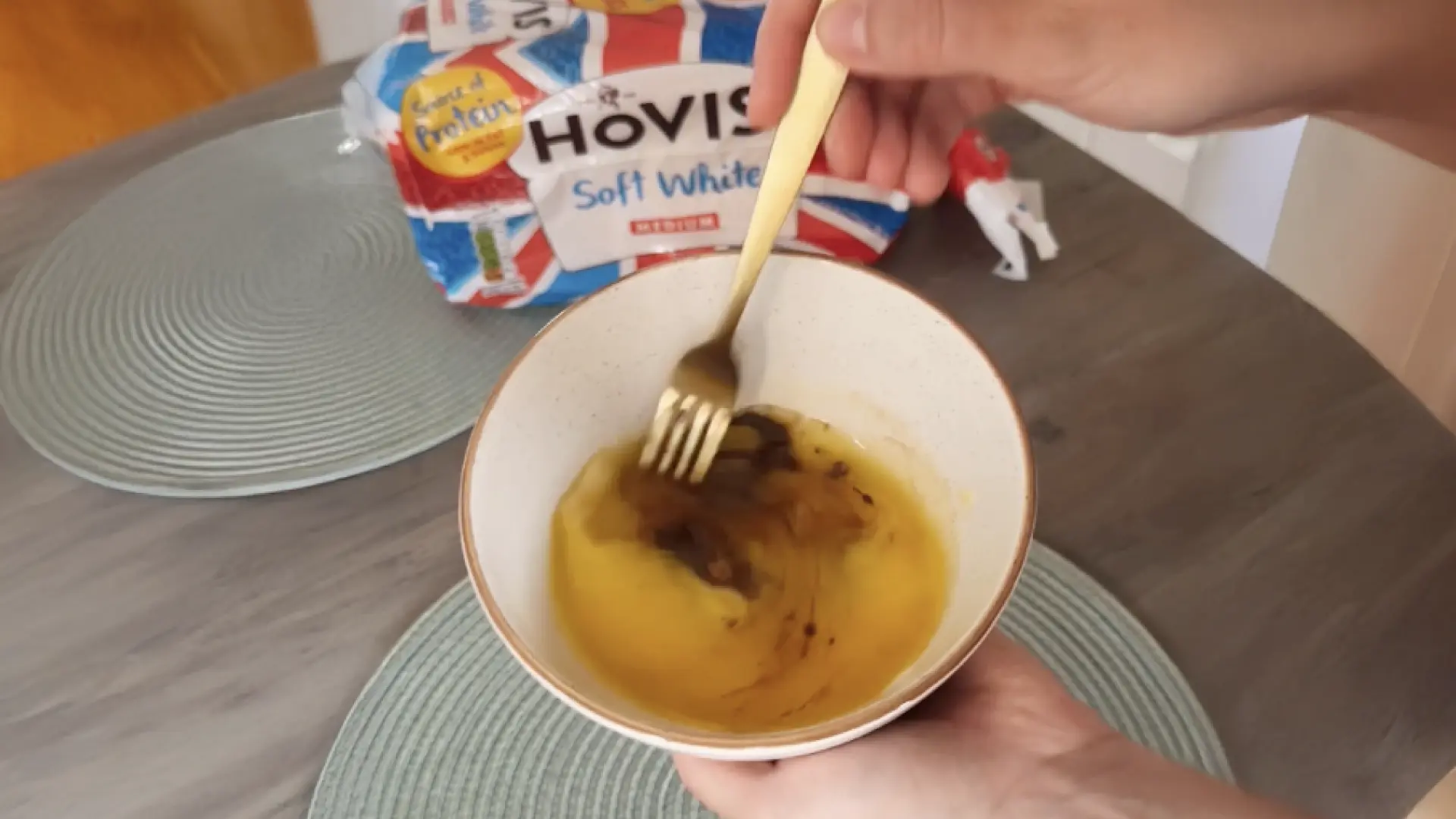 A hand using a gold fork to whisk liquid mixture in a bowl, with a bag of Hovis Soft White bread in the background.