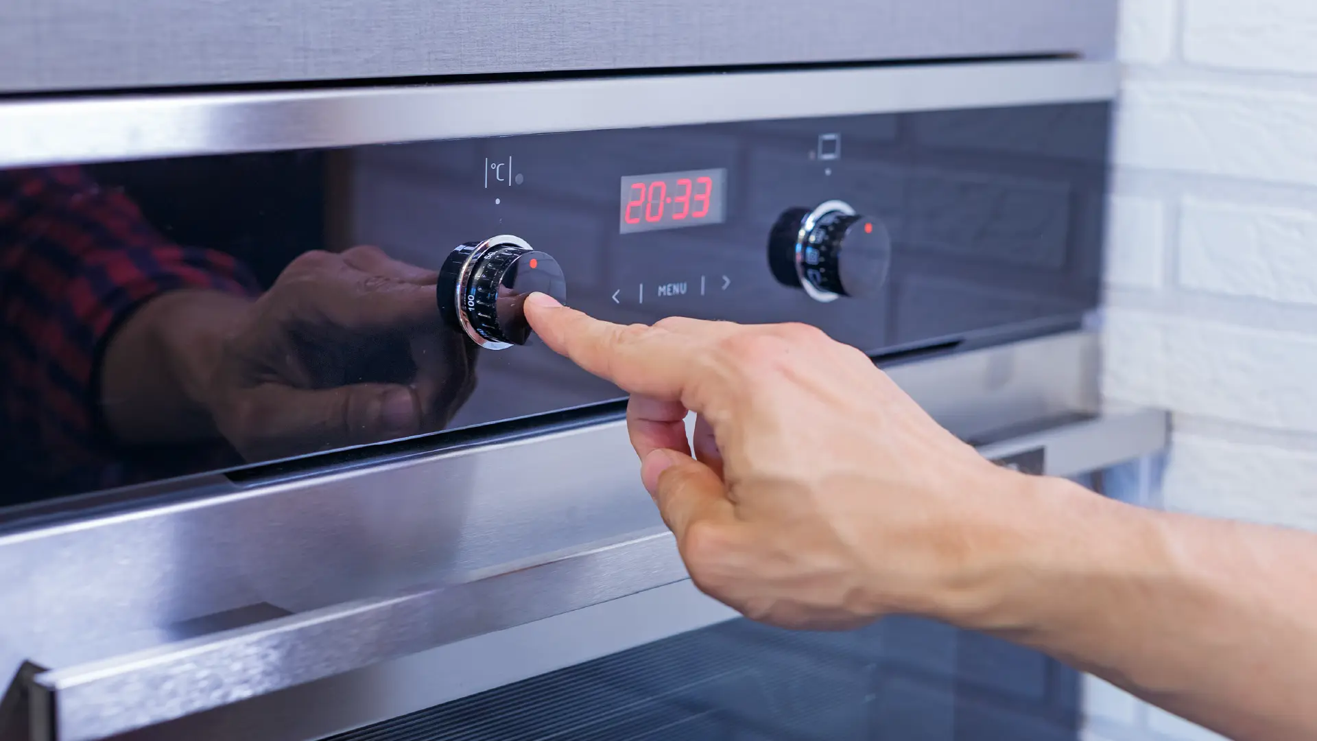 A person's hand adjusts the temperature dial on a modern stainless steel oven with a digital red display showing 20:33.