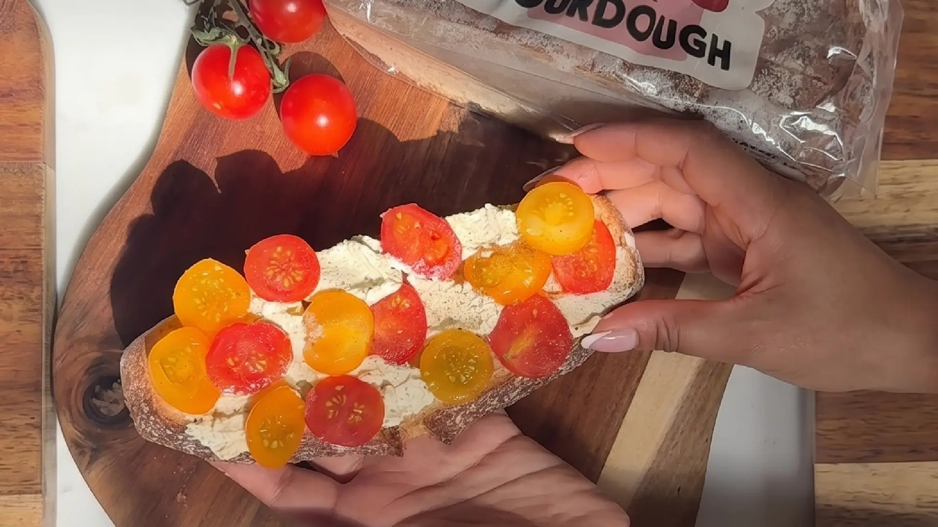 Hands holding a slice of sourdough toast slathered with goat cheese and topped with red and yellow cherry tomatoes.