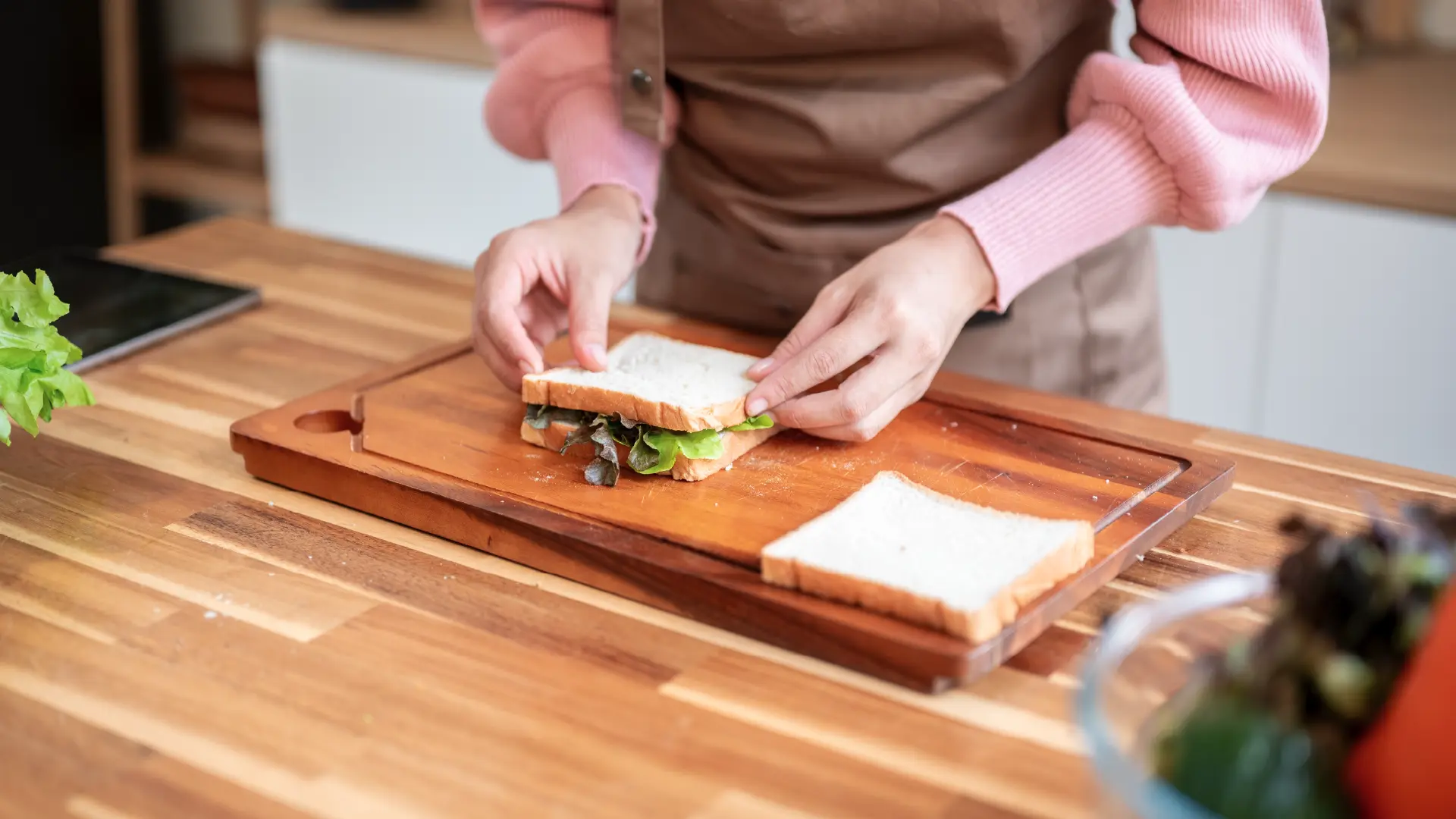 Person wearing an apron places the top slice of white bread onto a fresh vegetable sandwich on a wooden cutting board.