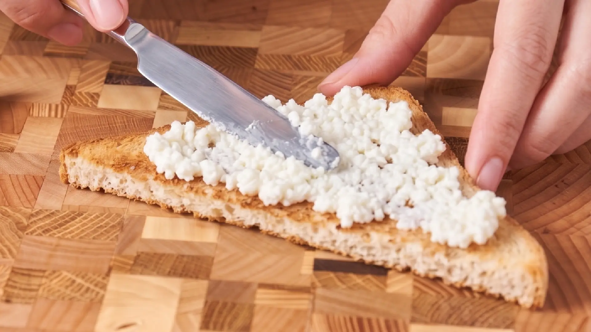 Close-up of hands using a metal knife to spread textured white ricotta cheese onto a triangular slice of golden toast.
