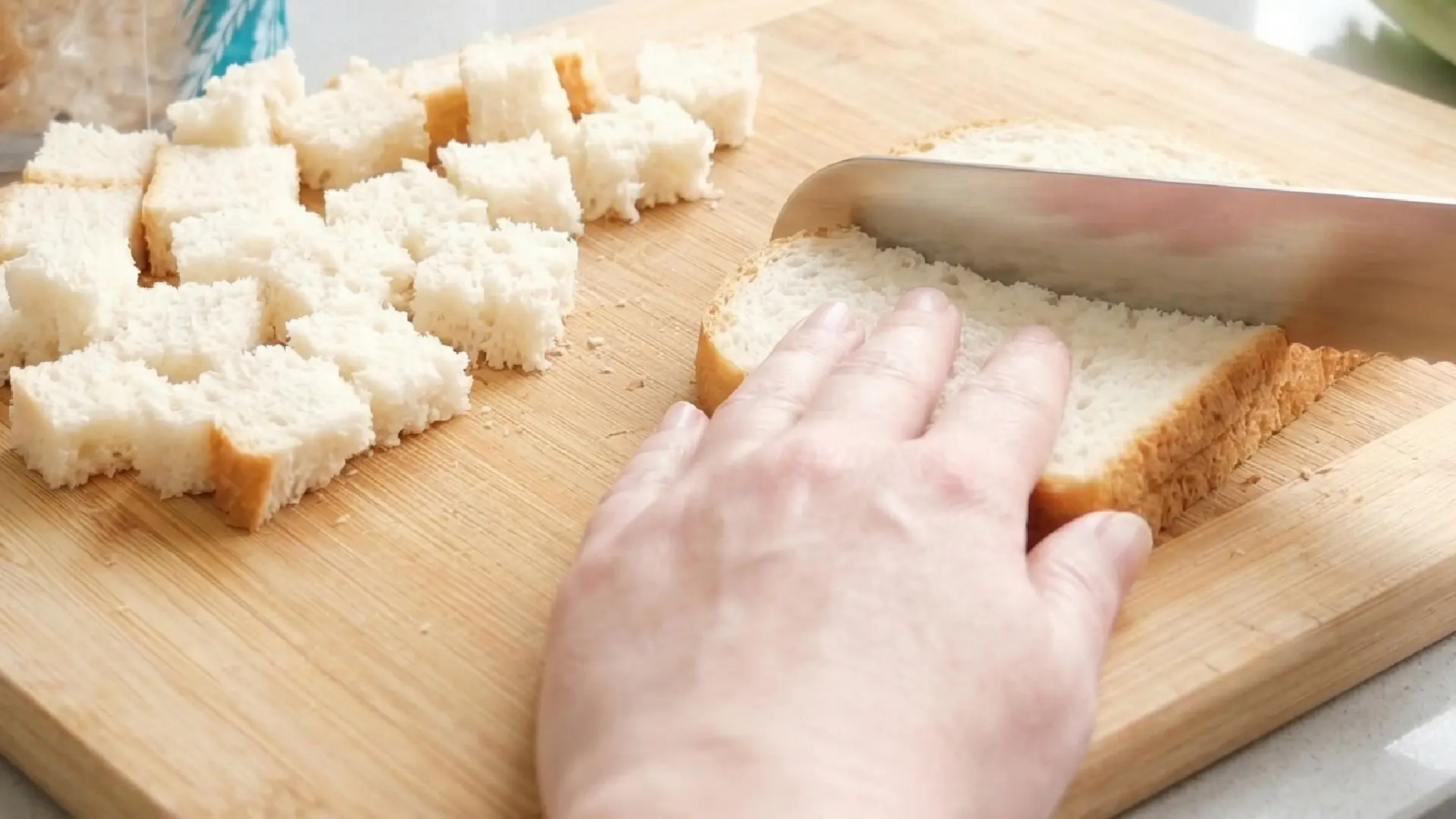 Close-up of a person slicing white bread into small cubes on a wooden cutting board to prepare homemade croutons.
