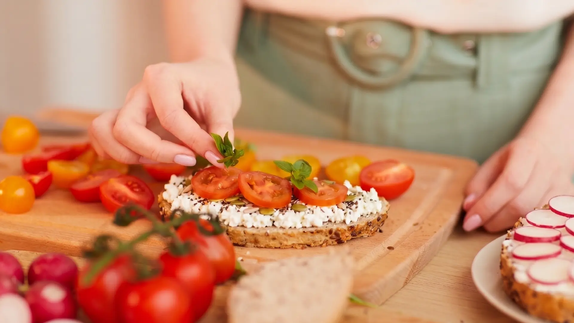 A person adds basil to ricotta toast with tomatoes on a wooden board, with radishes and cherry tomatoes nearby.