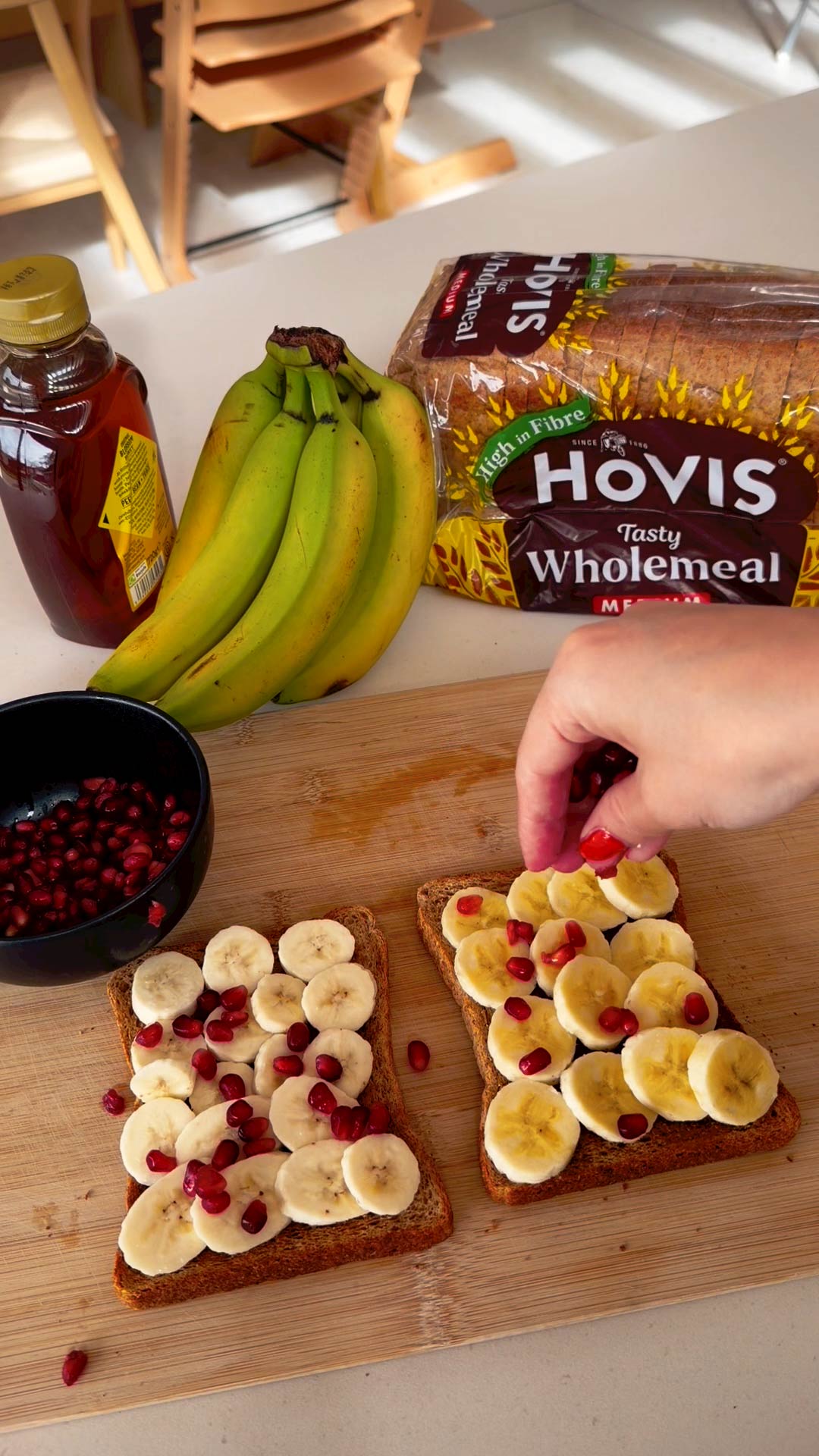 A high angle view showing the preparation of open faced toast topped with banana and pomegranate seeds on a wooden cutting board.