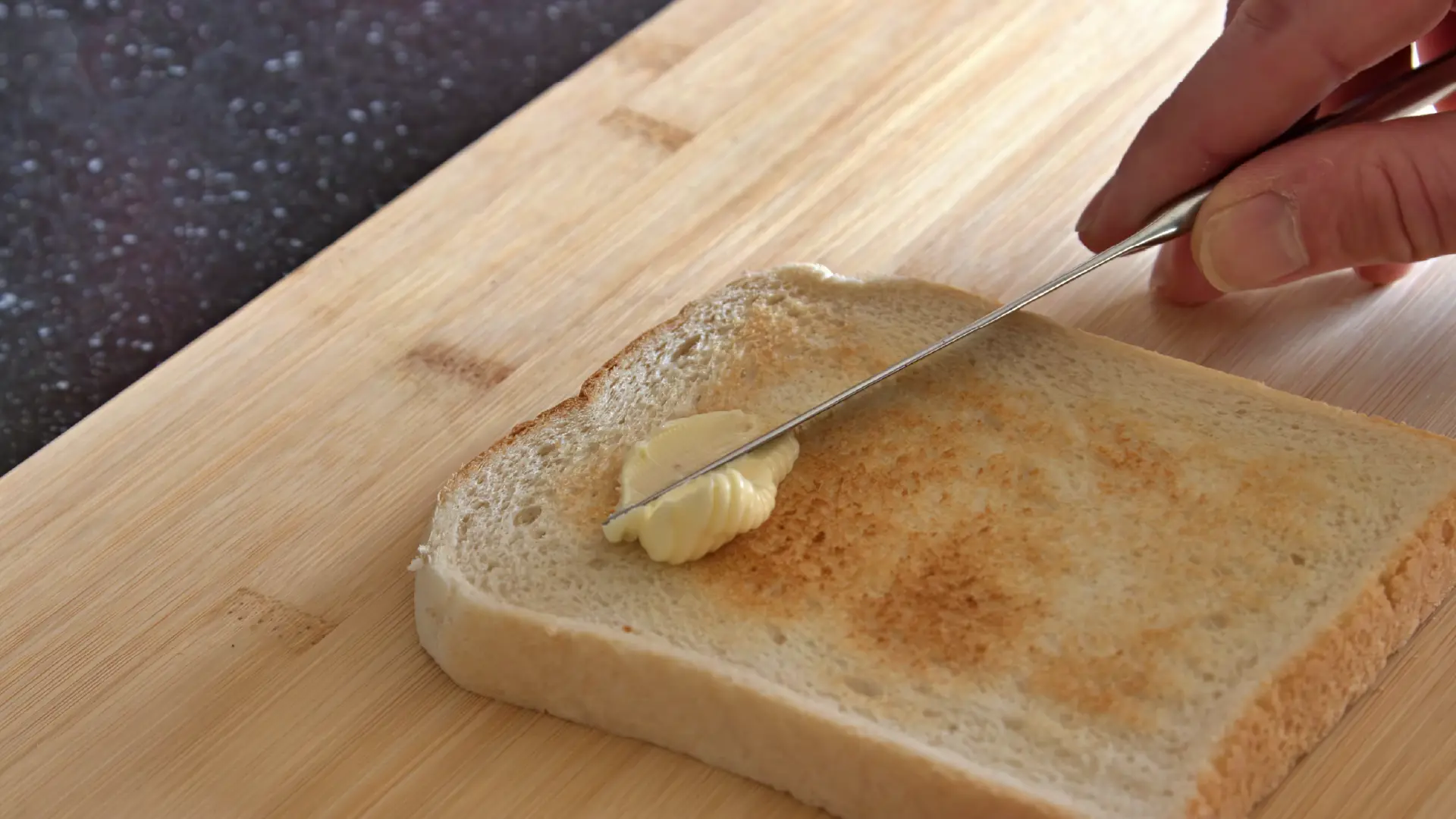 A hand uses a thin metal knife to spread a pat of yellow butter onto a slice of golden-brown toast on a wooden board.