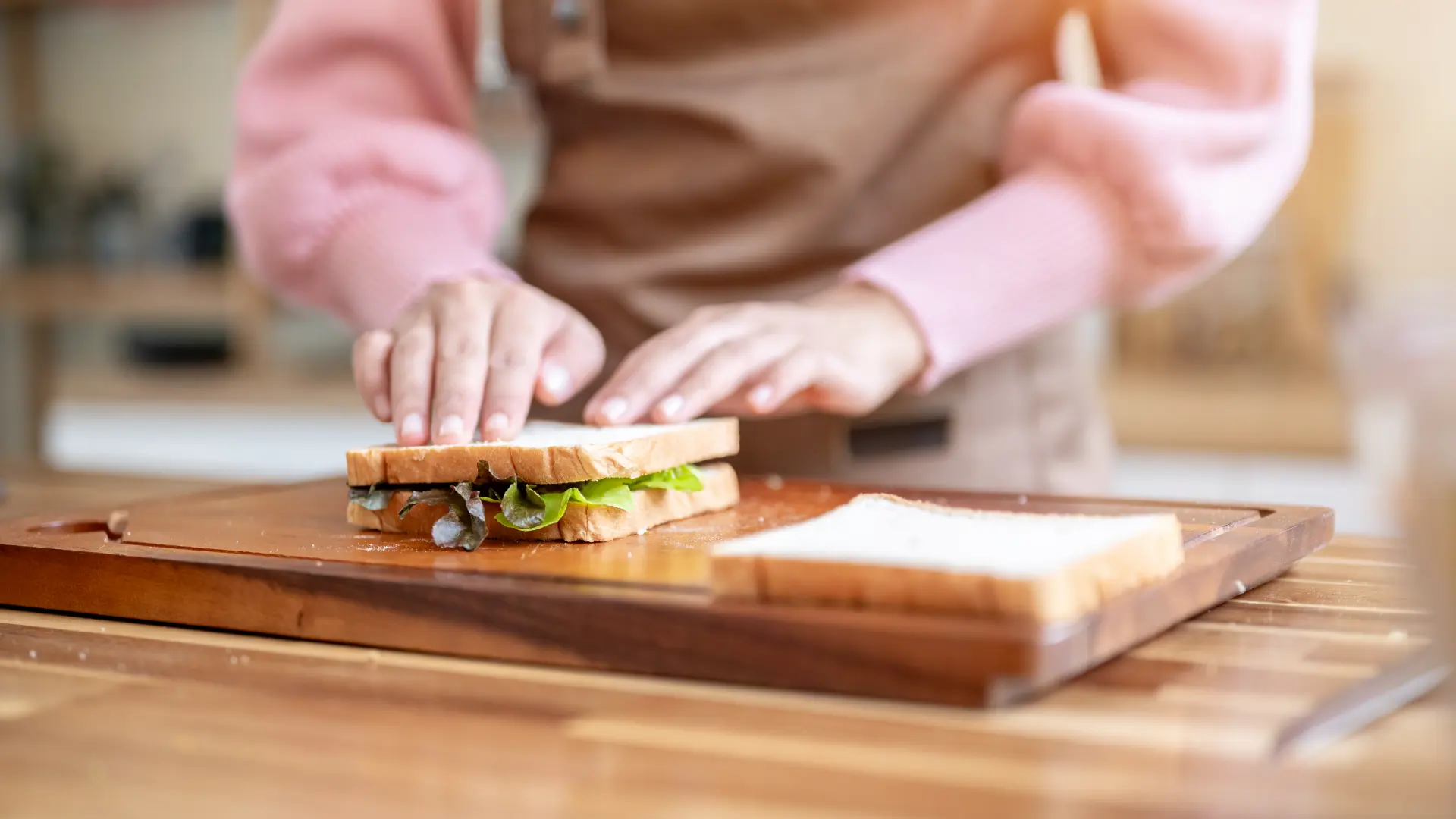 A person in a pink shirt pressing down on a lettuce sandwich on a wooden cutting board in a bright kitchen.