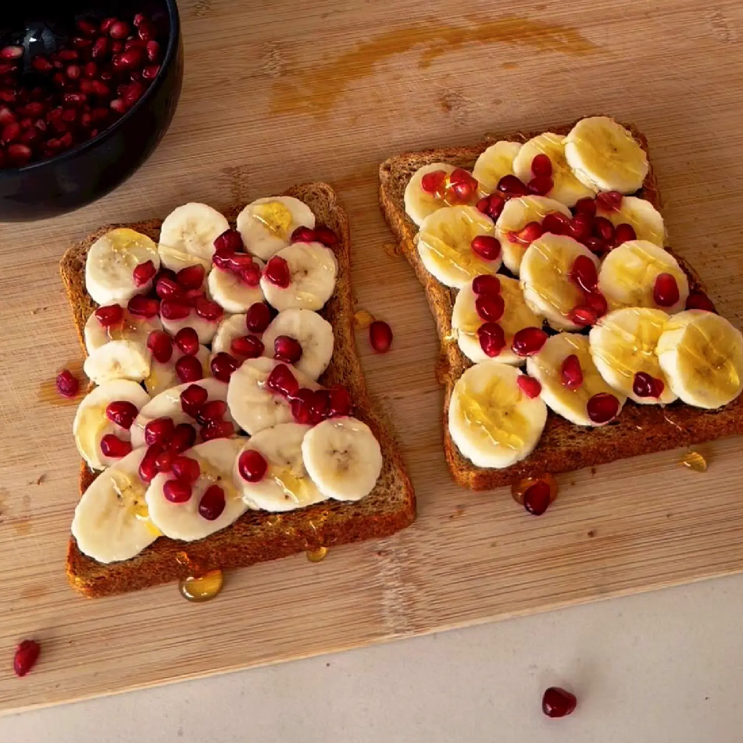 Two slices of wholemeal toast on a wooden board, topped with sliced bananas, pomegranate seeds and a drizzle of honey.