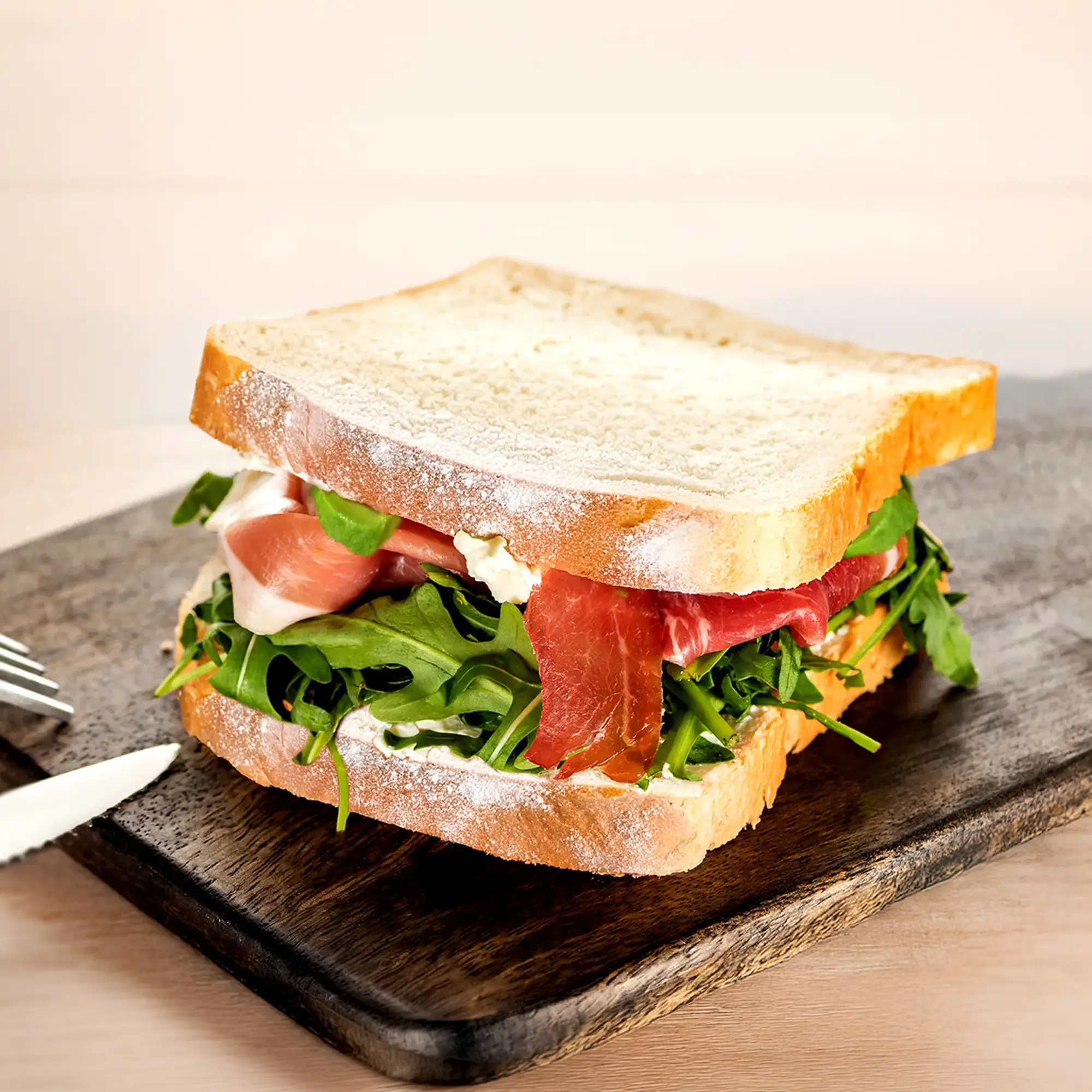 A sandwich made with Hovis Signature White bread sits on a dark wooden cutting board. The sandwich is filled with cheese spread, fresh rocket, and delicate slices of cured ham. The top slice of bread is slightly askew, revealing the delicious layers inside. A small portion of a knife and fork are visible to the left. 