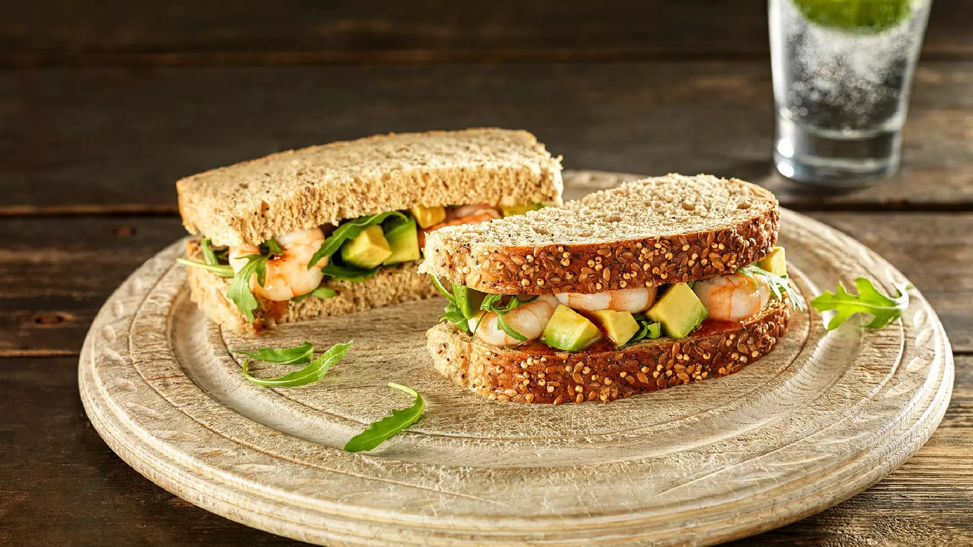 A Hovis Seeded Batch sandwich, halved and filled with prawns, cubed avocado, and rocket leaves, served on a clay plate.