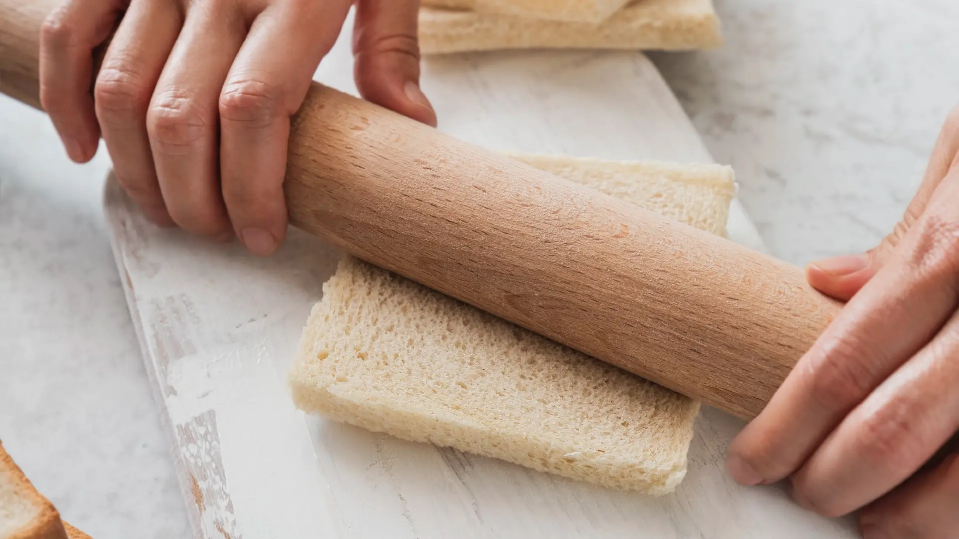 Hands using a wooden rolling pin to flatten a crustless slice of white bread on a marble cutting board.