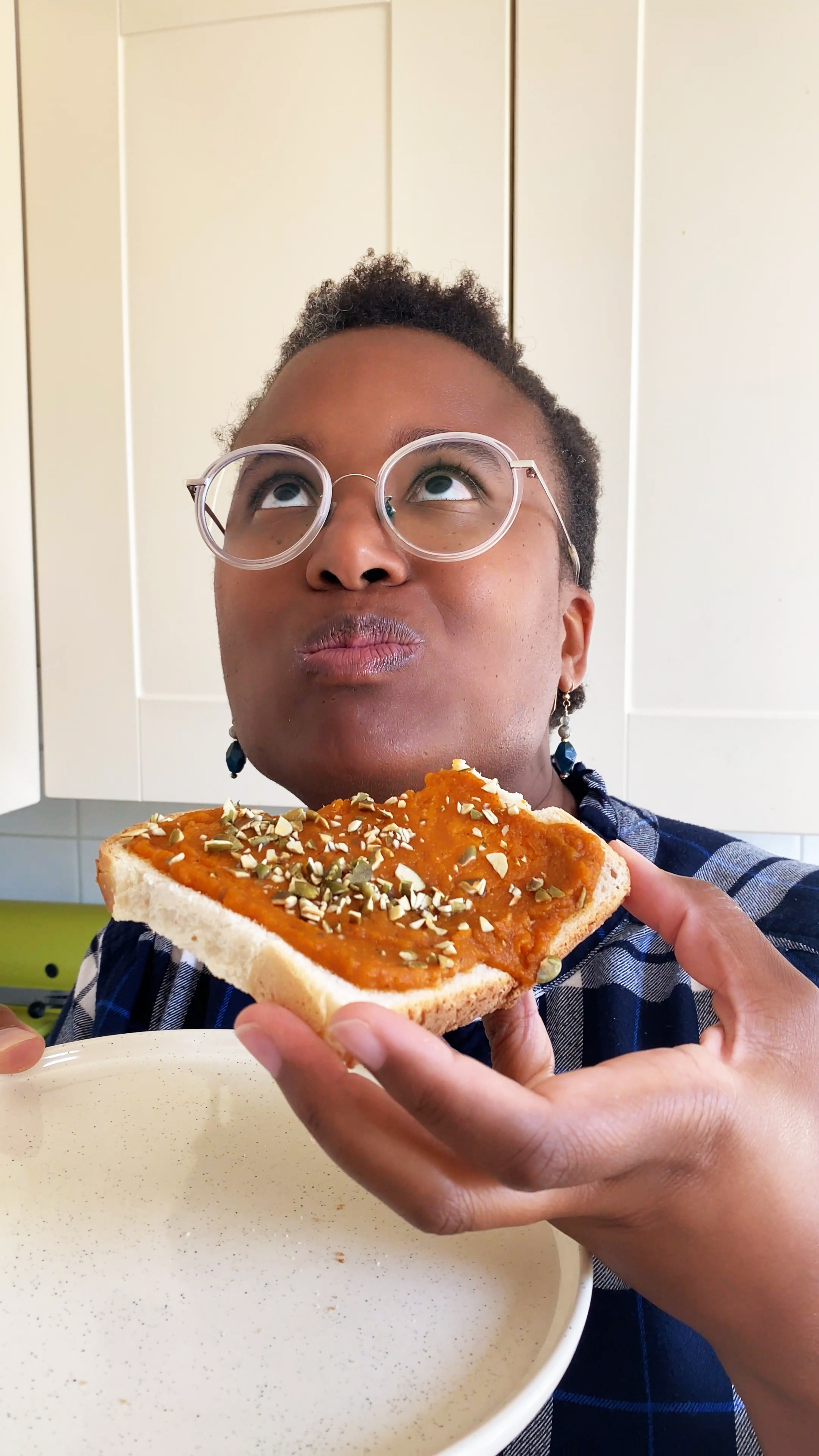 A person wearing glasses and a blue checkered shirt is enjoying a slice of toast topped with a generous layer of pumpkin-coloured spread and sprinkled with seeds. They hold the toast close to their mouth while looking playfully upward, cheeks slightly puffed as if savouring the bite. The background shows a bright kitchen, giving the moment a cosy, homemade feel.