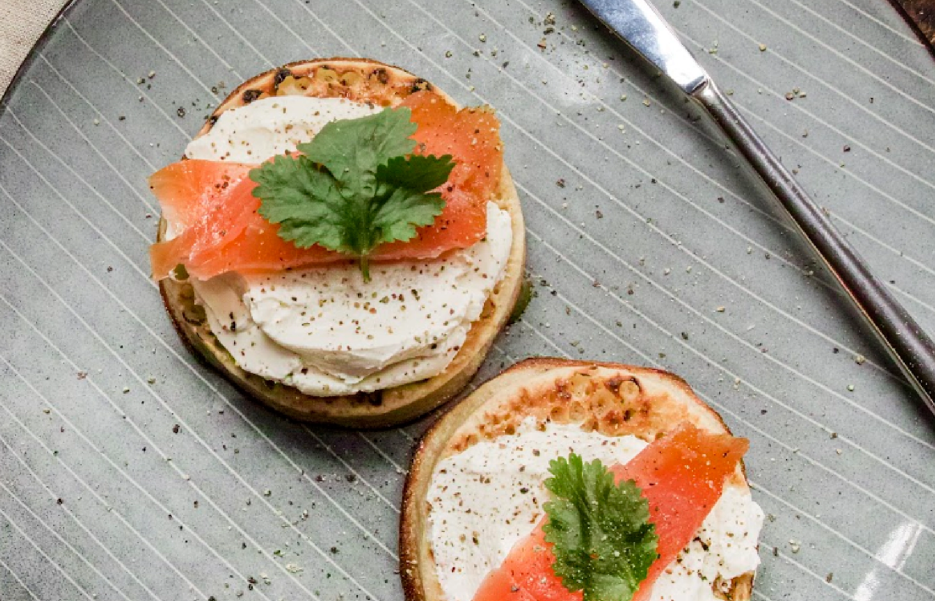 Top view of two slightly toasted Hovis Crumpets topped with cream cheese, a thin slice of smoked salmon, a parsley leaf, and ground pepper.