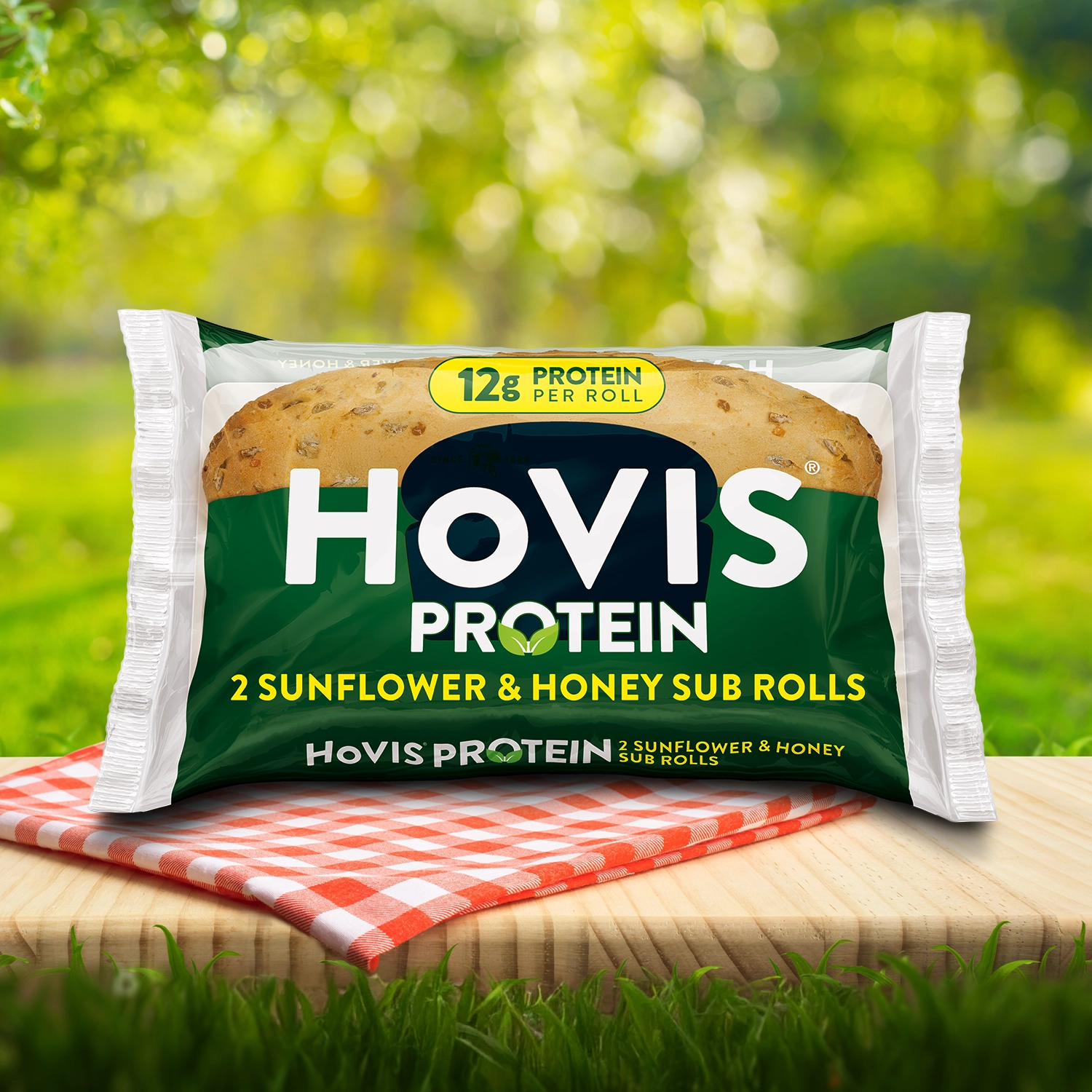 Hovis Protein Sunflower Honey Sub Rolls packaging on a picnic table with a green outdoor background. The pack shows two soft sub rolls with delicious sunflower seed on top. A red gingham cloth adds a picnic feel to the setting.