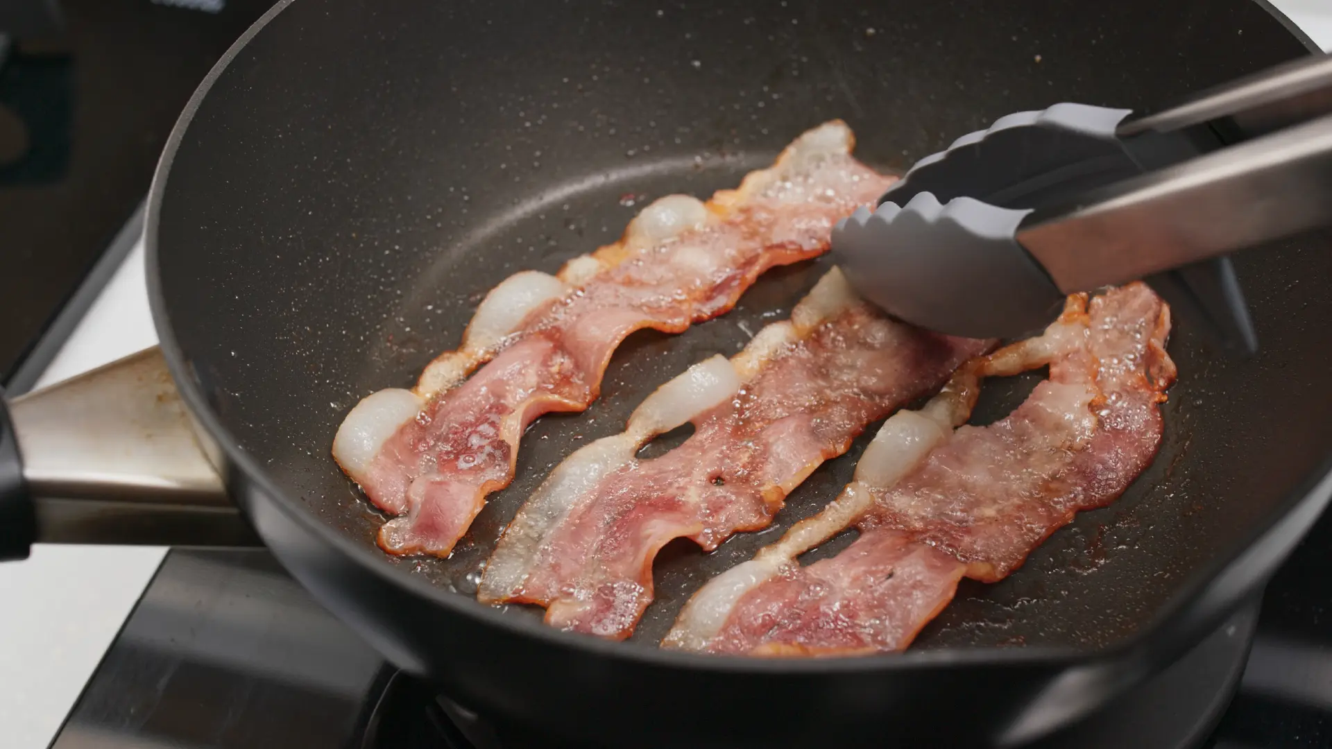 Three strips of bacon sizzle in a dark frying pan, being flipped with silver and grey kitchen tongs.