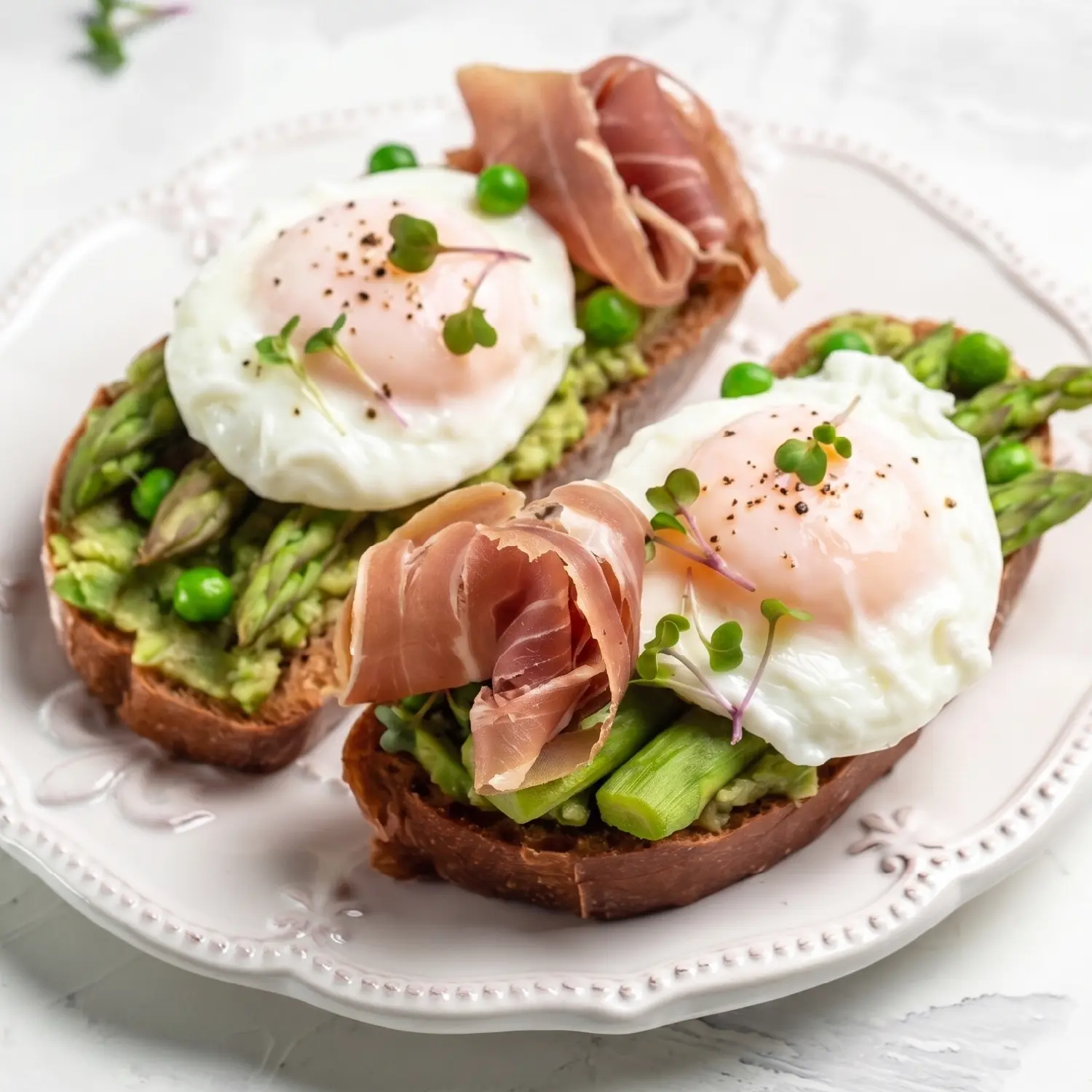 Two toasts with mashed avocado, asparagus, peas, prosciutto, and soft poached eggs topped with microgreens and pepper.