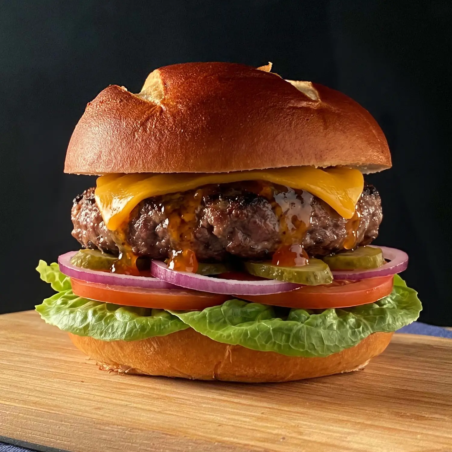 Gourmet cheeseburger with melted cheddar, lettuce, tomato, red onion, pickles and sauce on a toasted pretzel bun.