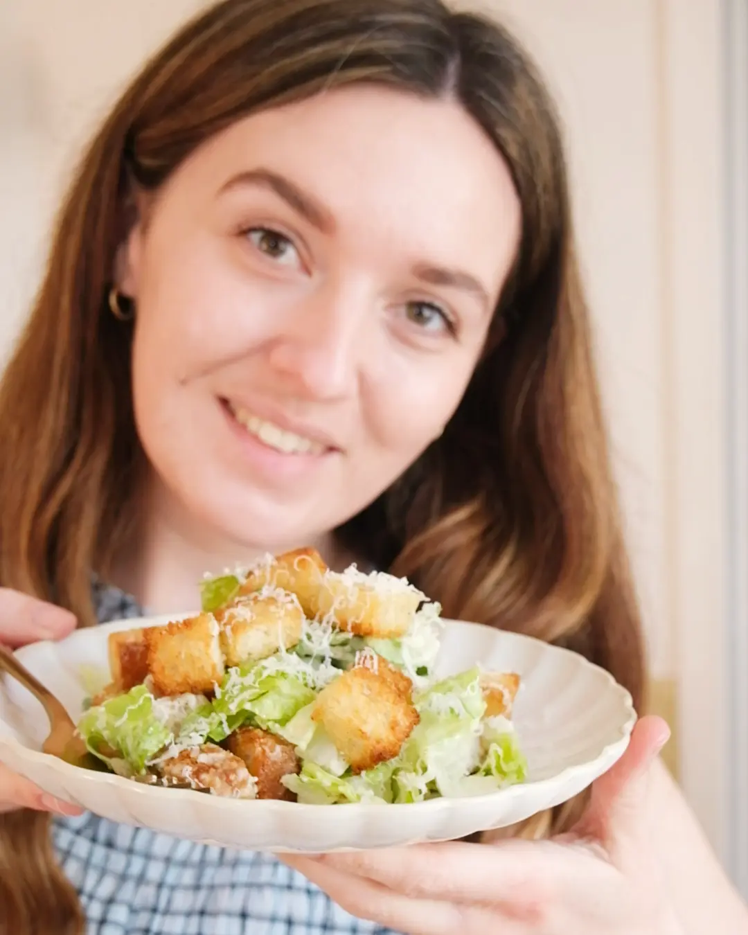 A woman smiling and holding a white wavy plate filled with a fresh Caesar salad topped with large, golden croutons.