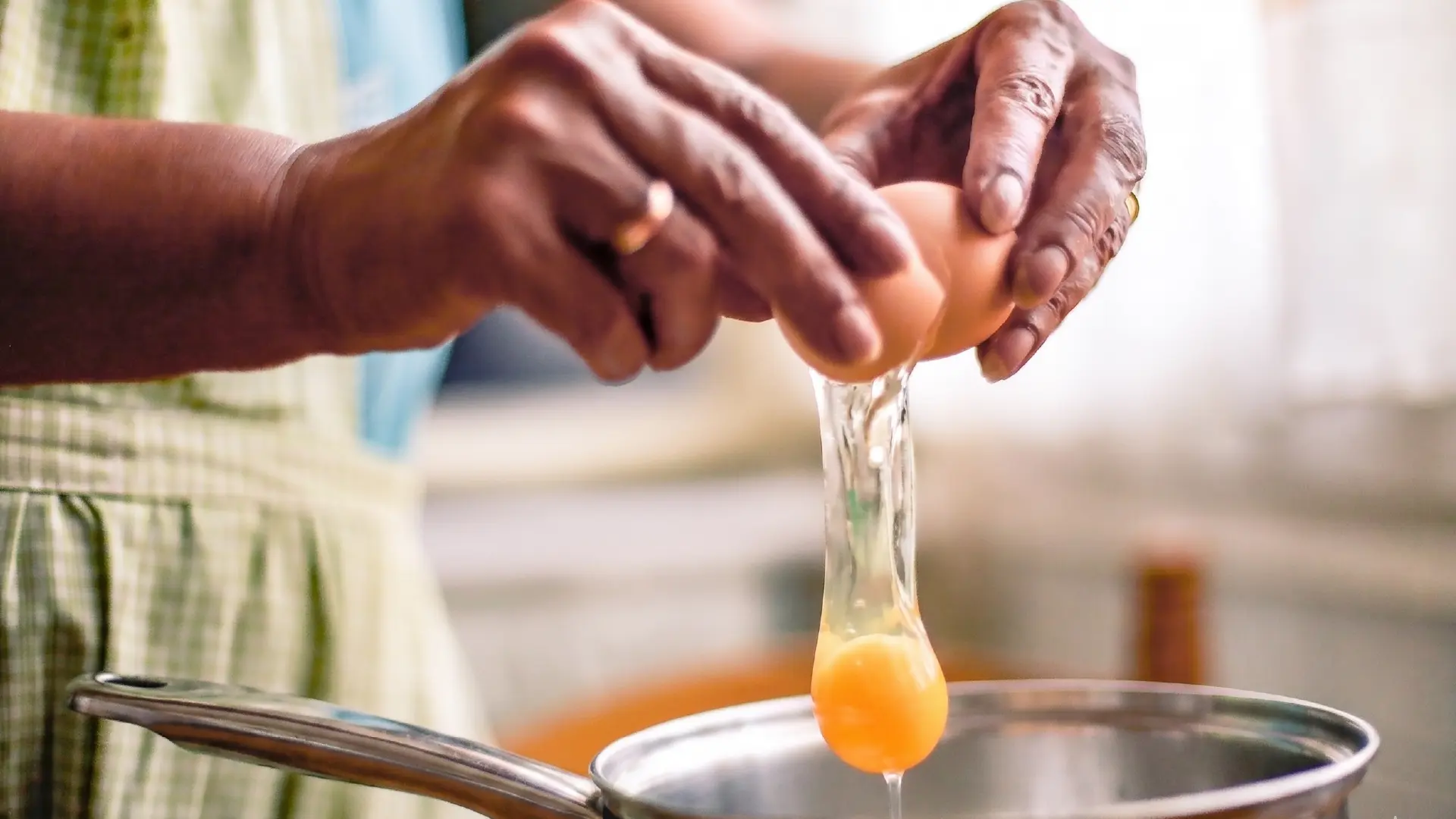 Close-up of hands cracking a brown egg into a stainless steel saucepan, with the yolk and white falling in mid-air.