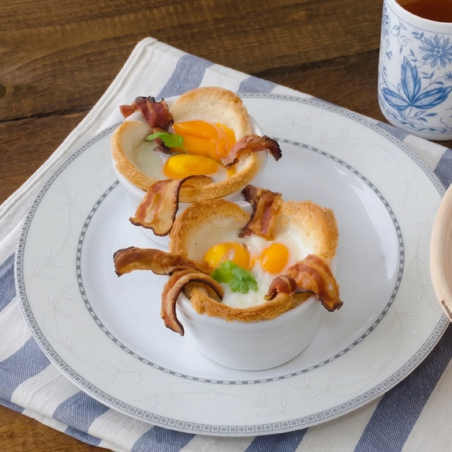 Two egg and bacon pastry cups served in a white ramekin on a patterned plate, garnished with fresh herbs.