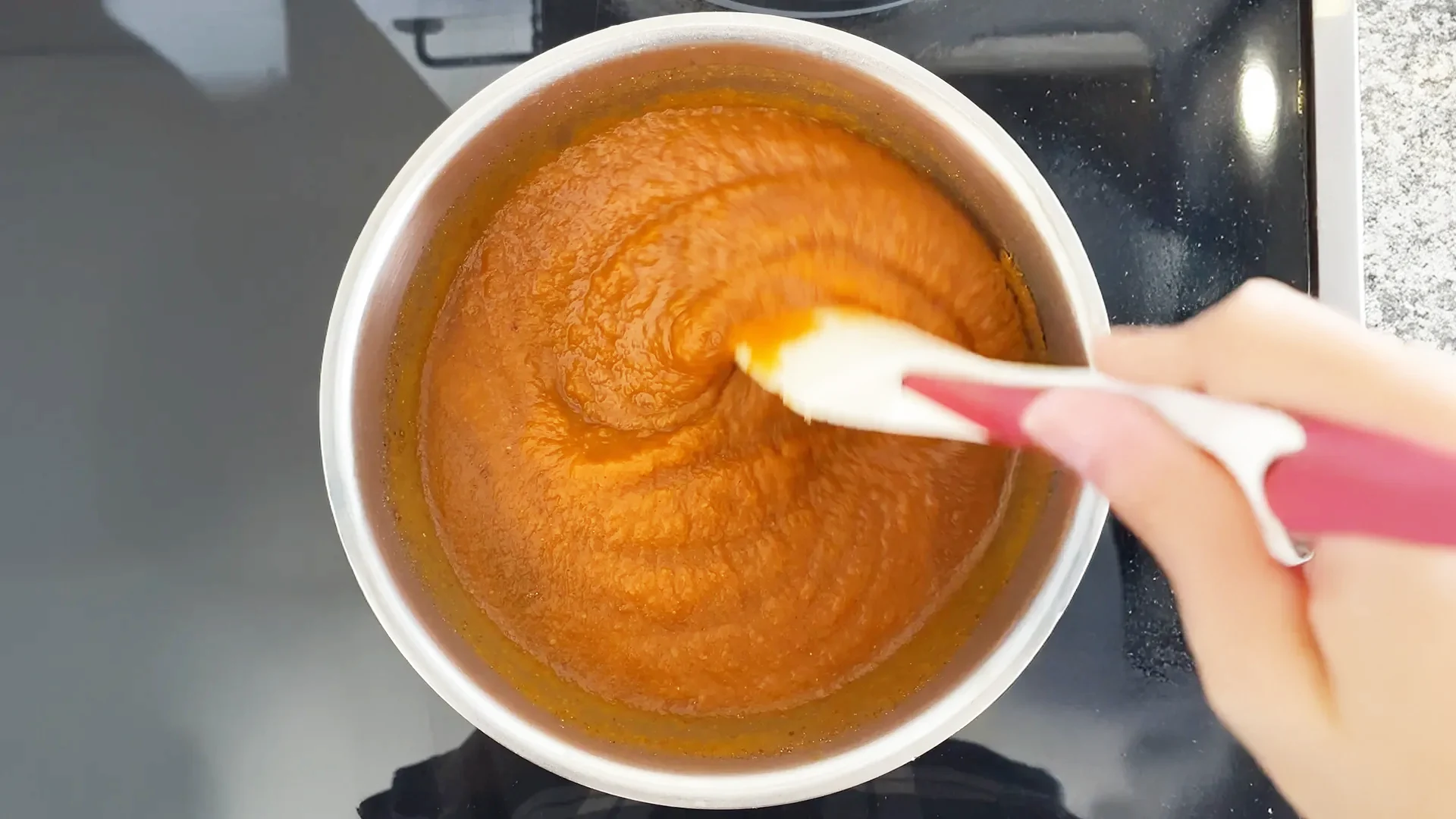 Someone is stirring the pumpkin butter mix on a hob with a pink and white spoon.