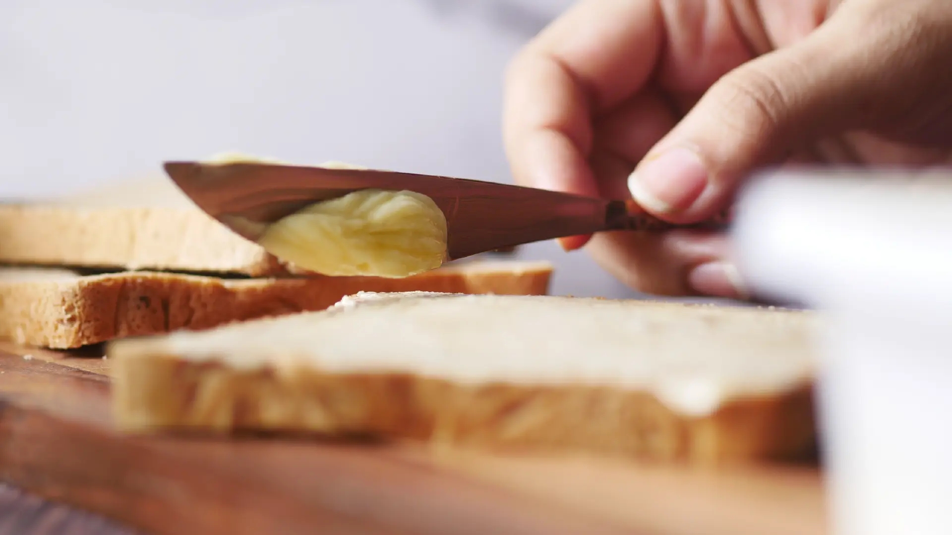 A hand using a butter knife to spread a creamy layer of mayonnaise or butter onto a slice of white bread.