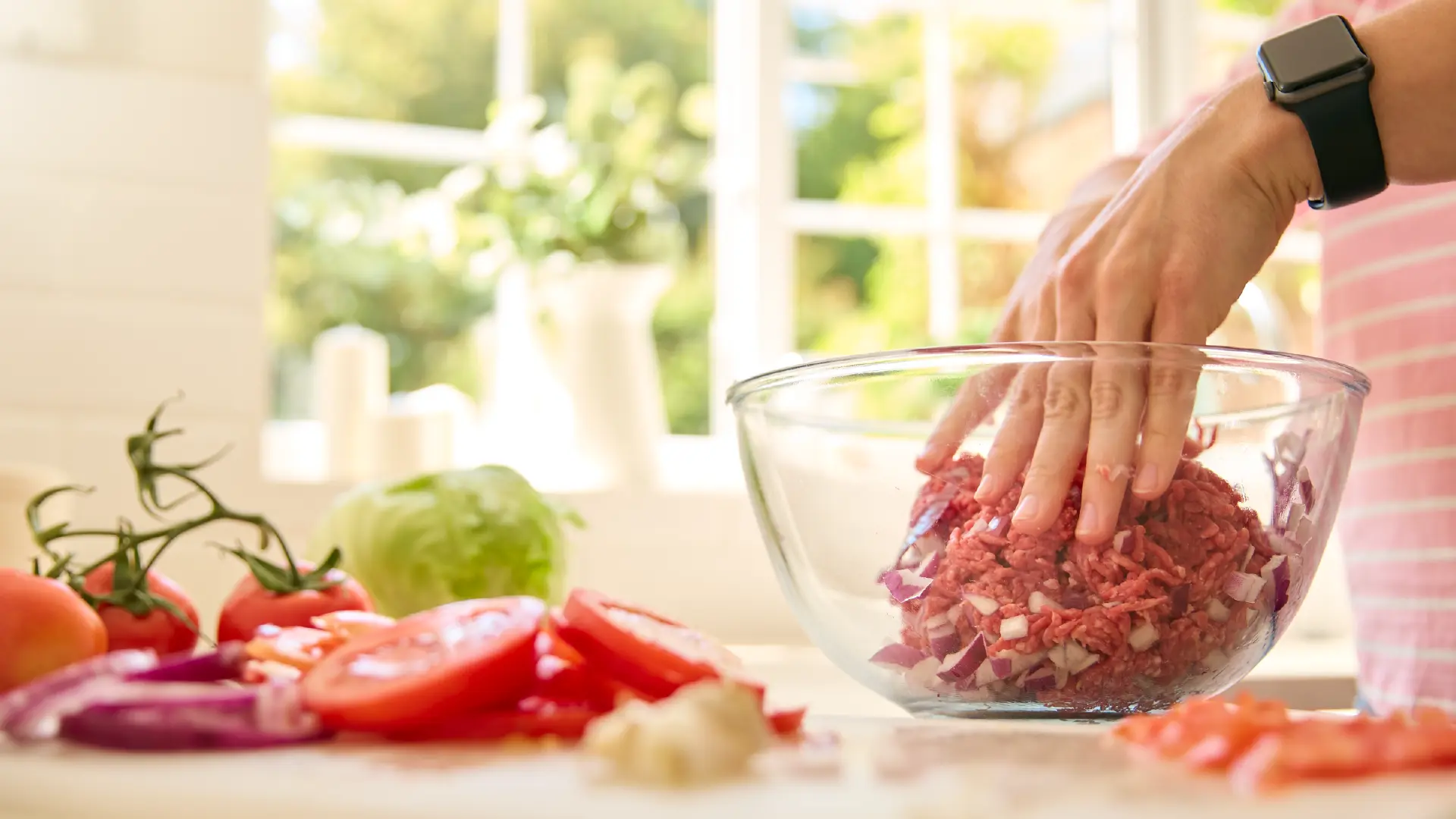 Hands mixing lean beef mince and onions in a glass bowl, with fresh tomatoes and lettuce ready on a bright kitchen counter.