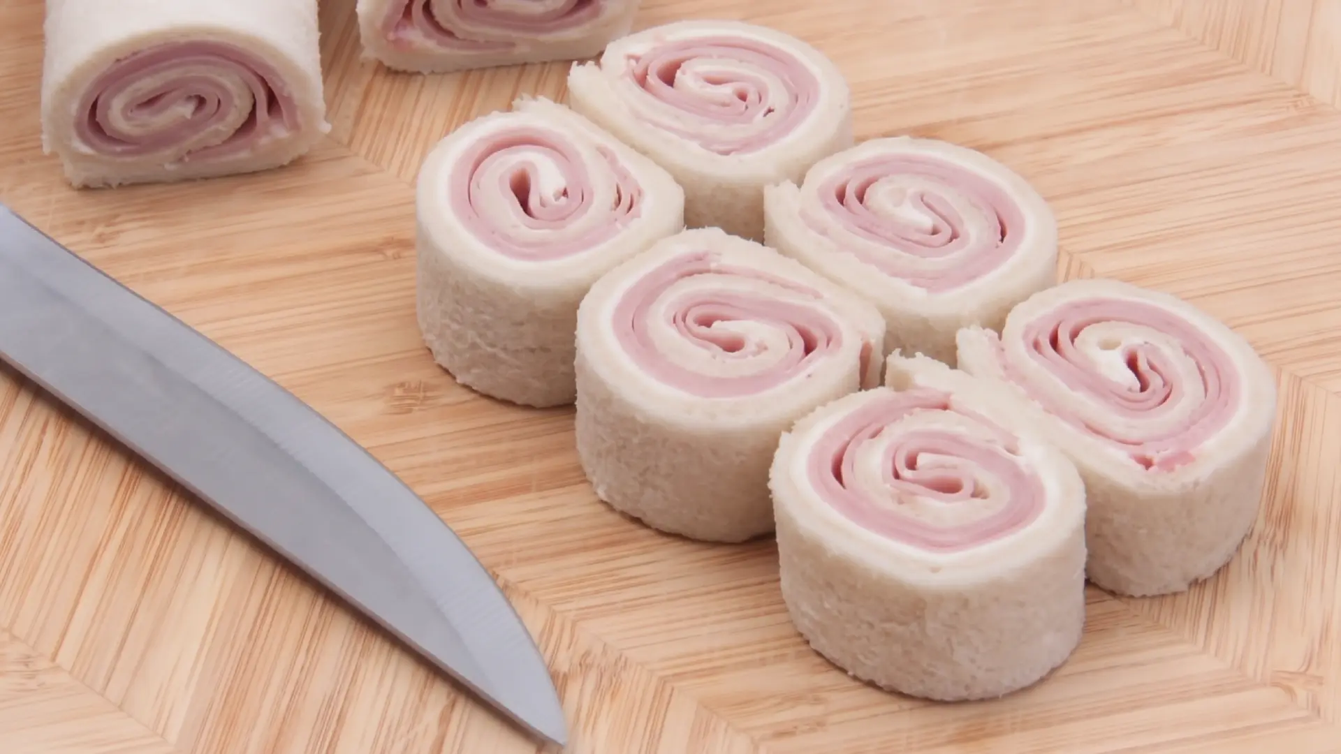 Sliced pinwheel sandwiches made of white bread and ham sitting on a light wooden cutting board next to a chef's knife.