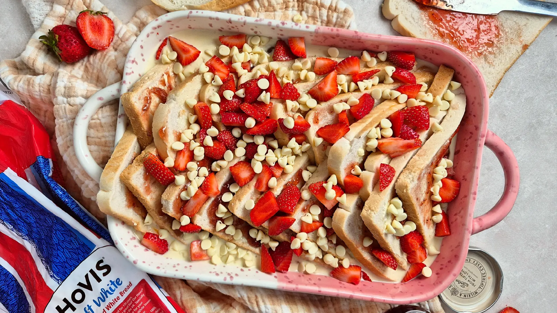 A pink baking dish filled with bread pudding topped with white chocolate chips and fresh sliced strawberries.
