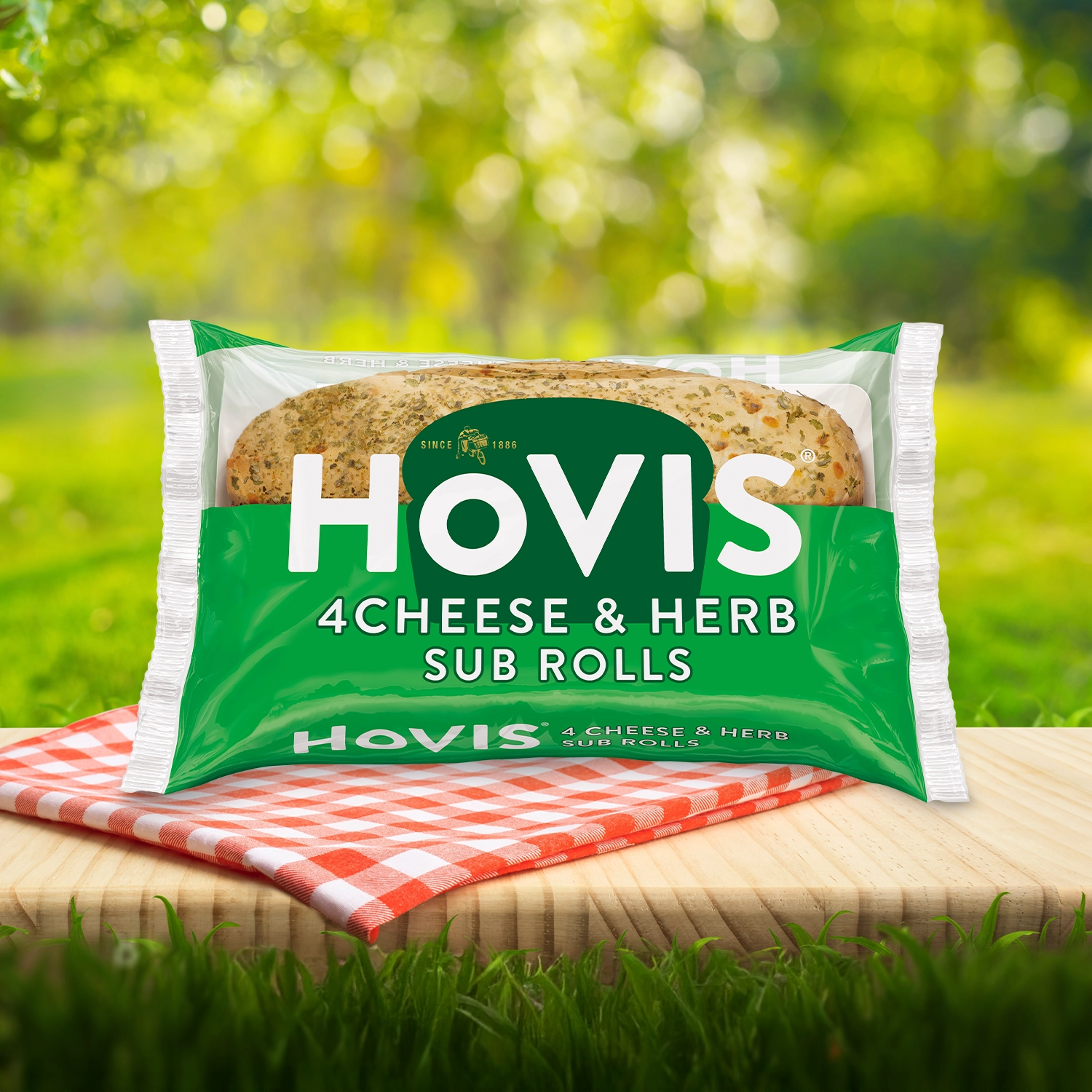 Hovis Cheese & Herb Sub Rolls Sub Rolls packaging on a picnic table with a green outdoor background. The pack shows two soft sub rolls with herbs and highlights “Deliciously soft fusion of cheese and herbs.”  A red gingham cloth adds a picnic feel to the setting.